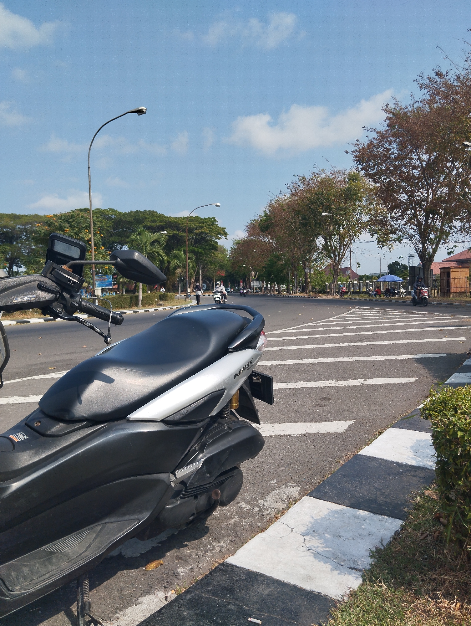 A motorcycle is parked on the side of a road with a pedestrian crossing in the foreground. The road is lined with trees and streetlights. In the background, there are buildings and people walking. The sky is blue with some clouds. The scene is likely in Betro, Indonesia, given the context.