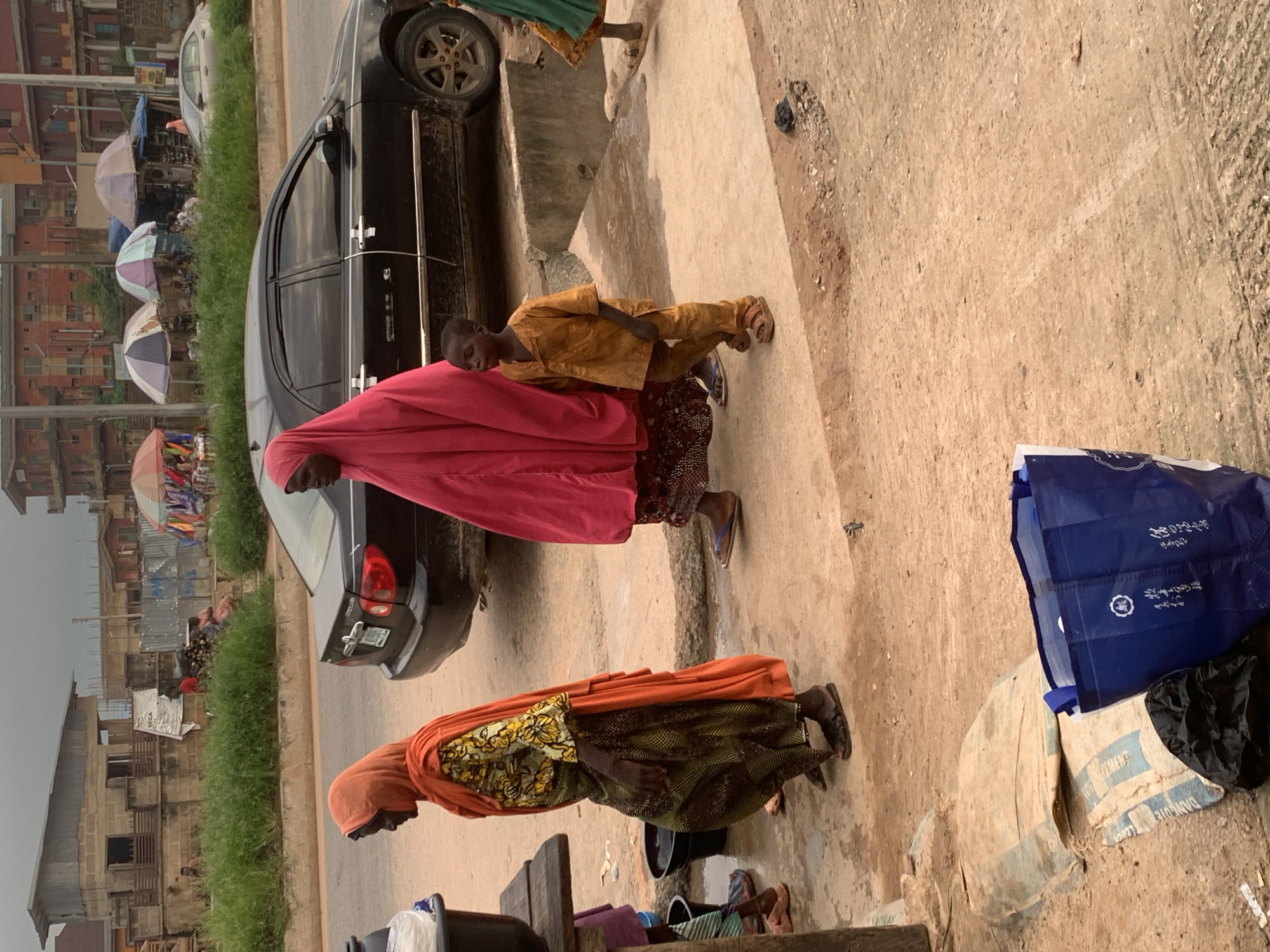 In Shagamu, Nigeria, a street scene unfolds with two adult women and a young boy walking alongside a black sedan. The woman on the left wears an orange headscarf and a patterned dress, while the woman in the center is clad in a vibrant pink abaya. The young boy, dressed in a simple gold outfit, walks between them. They are on a dirt and concrete path next to a road, with a black car parked on the right. In the background, buildings and market stalls with umbrellas are visible. Bags, including a blue reusable shopping bag, are scattered on the ground, suggesting a busy marketplace or residential area.