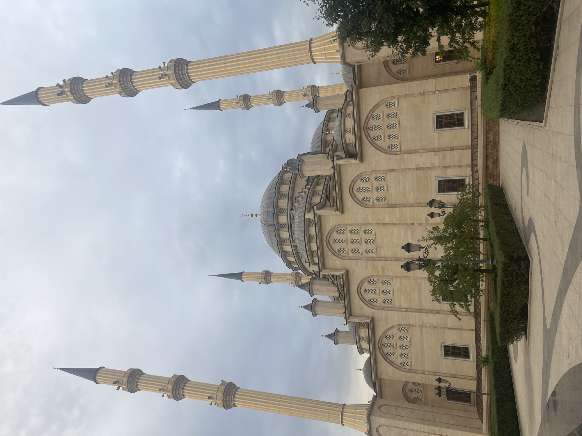 This image shows the Akhmad Kadyrov Mosque in Grozny, Russia, on an overcast day. The mosque is a large, ornate building made of pale stone with multiple domes and minarets. The central dome is large and rounded, topped with a crescent moon. Four tall, slender minarets with pointed dark spires flank the mosque, two on each side. These minarets are decorated with vertical fluted columns and balconies. The facade of the mosque features numerous arched windows with decorative grills. In the foreground, a paved walkway with curved dark lines winds through manicured green hedges and a few small trees. Several ornate lampposts are also visible along the walkway. The sky is a muted gray with clouds, and there is a tree on the right side of the image. There are no people visible in the scene.