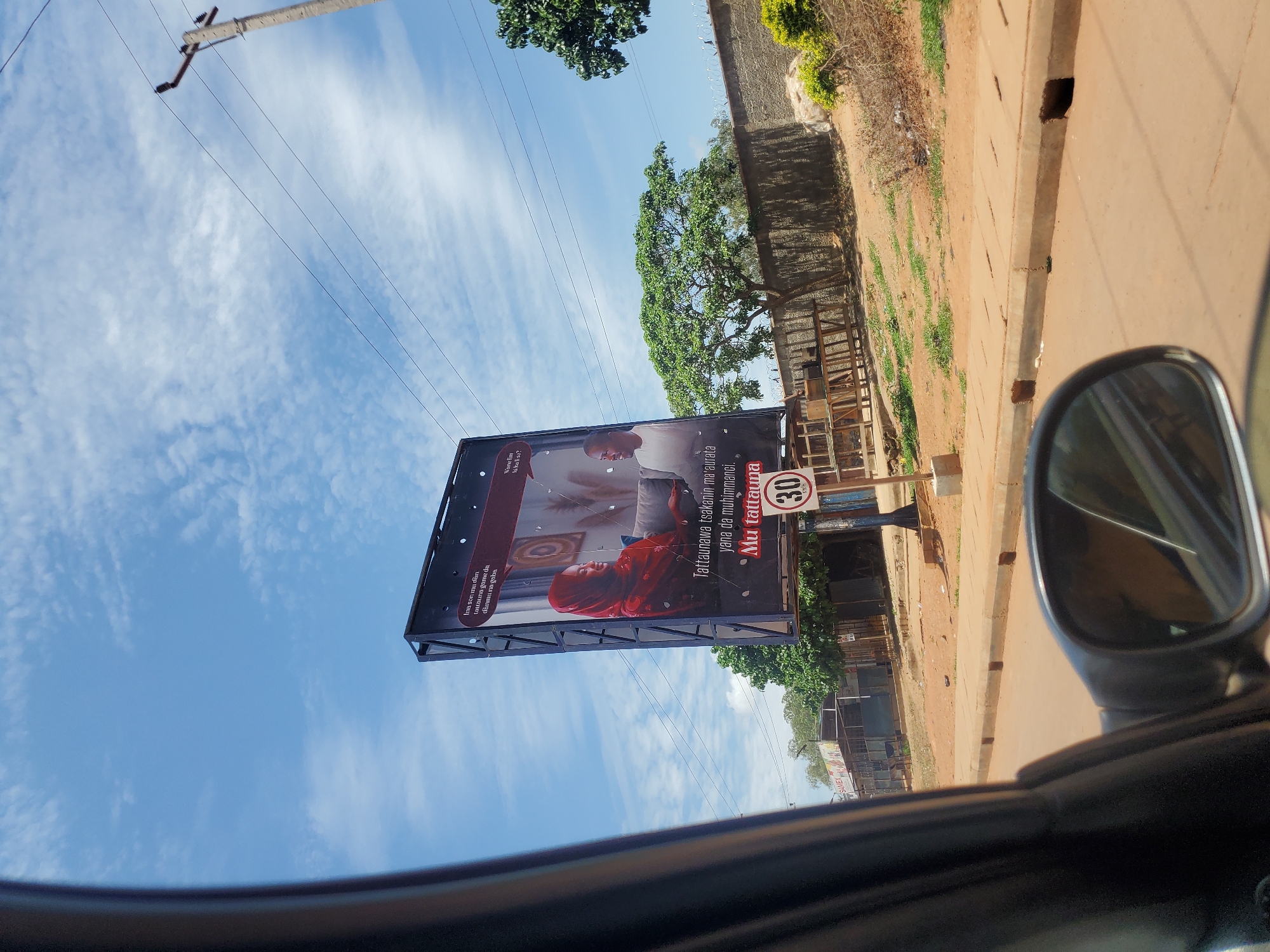 The image depicts a roadside scene in Television, Nigeria, captured from inside a vehicle, with portions of the car's dark interior and side mirror visible in the foreground. The sky is bright blue with scattered white clouds.
A large, double-sided digital billboard stands prominently on the left side of the road. The visible side displays an advertisement featuring two individuals: a woman in a red hijab and patterned top, seated on a light-colored sofa while looking at an unseen device; and a man in a white t-shirt, also seated on a sofa, looking towards the woman. The advertisement contains text in Hausa, which translates to: "I want us to discuss tomorrow's worries/concerns" (top left), "Discussion between spouses is important" (bottom center), and "Let's discuss" (bottom center). The MTN logo is partially visible in the top right corner of the billboard.
Adjacent to the billboard, a circular speed limit sign indicates "30 KM/H". The immediate road surface appears to be packed reddish-brown dirt, transitioning to a paved asphalt road beyond. A concrete sidewalk with multiple rectangular drainage openings runs along the right side of the dirt road.
In the background on the left, several low-rise commercial structures with corrugated metal roofs are visible, one sign partially displaying "SAVEX". On the right side, beyond the road and sidewalk, a concrete wall topped with barbed wire runs horizontally, backed by various trees and sparse green vegetation. A concrete utility pole with power lines extends into the sky on the far right. The overall lighting suggests bright daylight conditions.