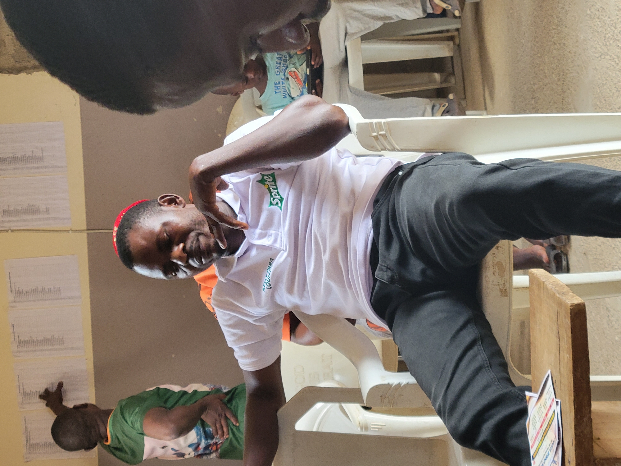 A man in a white Sprite-branded shirt and black pants sits in a white plastic chair in an indoor setting, possibly a room in Madalla, Nigeria. He is making a hand gesture with his right hand. The background includes walls with papers attached, and other people are present, sitting in chairs. The scene appears to be a casual meeting or gathering.