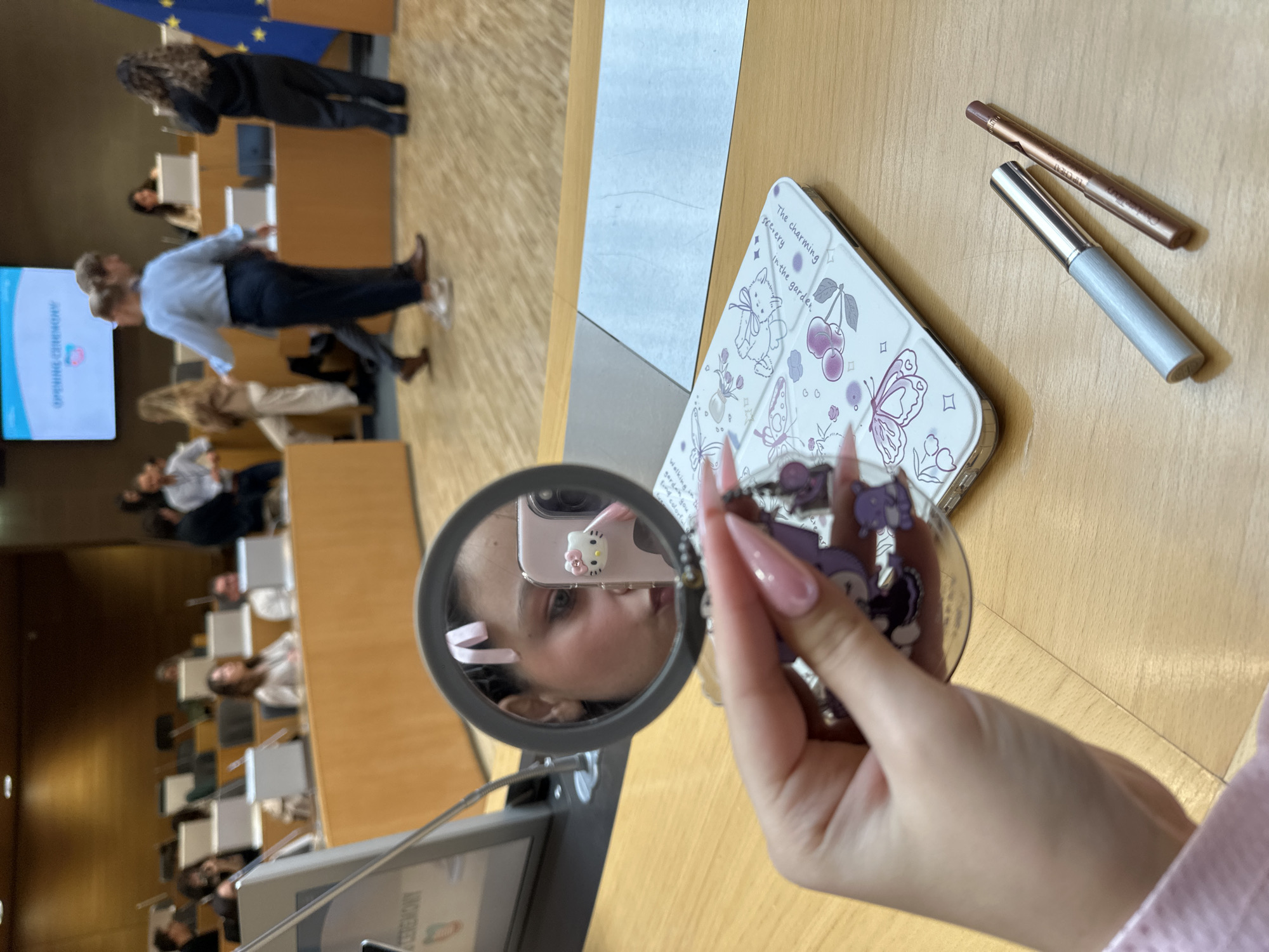 The image shows a woman sitting in an auditorium-style room that appears to be in session. Many people can be seen sitting at tables. A screen in the background displays the words "Opening Ceremony" in English. In the foreground, a woman is holding a round mirror, reflecting her face and the back of a cell phone with a "Hello Kitty" decoration. The table she is sitting at also contains a phone in a white case with cartoon designs, and two make-up pencils. The image was taken in Strasbourg, France.