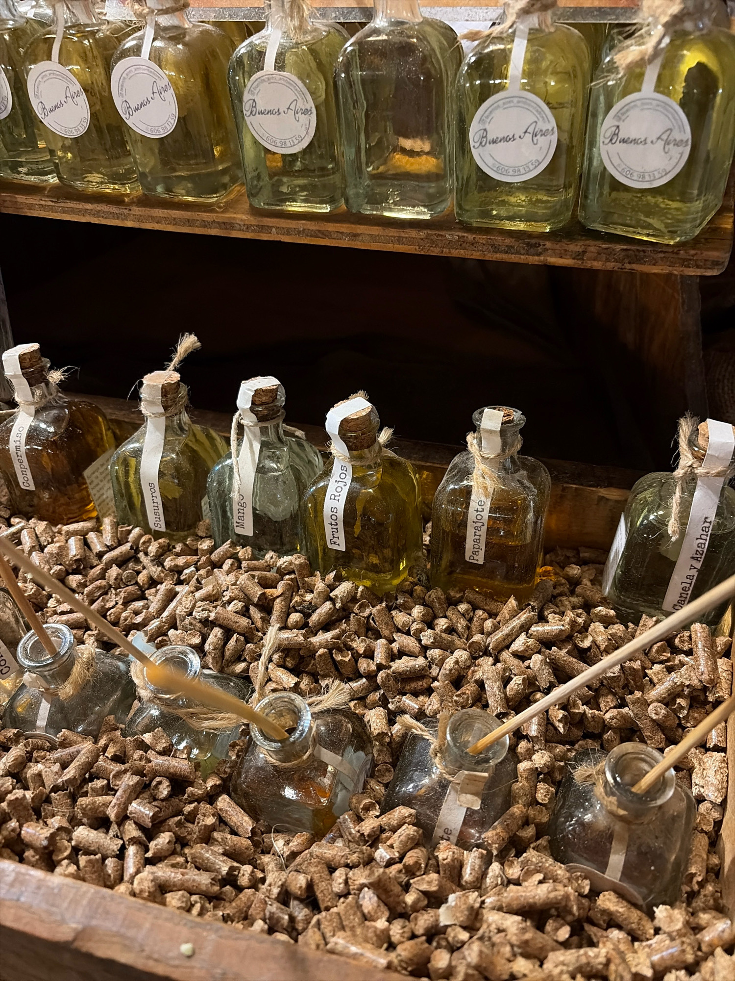 The image displays a collection of small glass bottles, some containing amber-colored liquid, arranged on a wooden shelf and in a wooden crate filled with wood pellets. The bottles on the shelf have circular white labels with the text "Buenos Aires" and a phone number. The bottles in the crate have paper labels with handwritten text, including "Conpermiso," "Susurros," "Mango," "Frutos Rojos," "Paparajote," and "Canela y Azahar." Several bottles in the crate have thin wooden sticks inserted into them, suggesting they might be diffusers or incense. The lighting suggests an indoor setting, likely a retail environment. The context of "Buenos Aires" on the labels, combined with the presentation of artisanal products, implies a shop or market stall. However, the image itself does not provide any specific visual cues to pinpoint the location within the city of Salou, Spain.