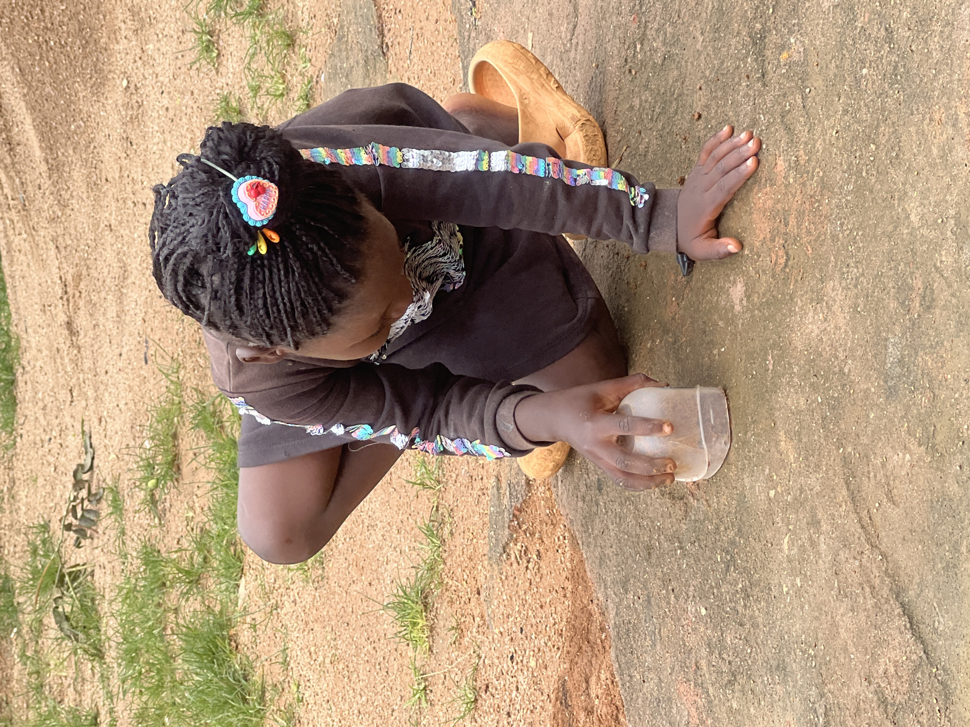 A young Black girl with braided hair, adorned with a heart-shaped hair clip, kneels on a concrete surface. She wears a dark brown sweatshirt with colorful sequin embellishments on the sleeves and a dark skirt. Her right hand holds a clear plastic container partially filled with dirt, while her left hand rests on the ground. She is wearing a pair of tan clogs. The background consists of dry, reddish-brown earth, patches of dry grass, and some scattered debris. The scene appears to be outdoors, in a somewhat unkempt, possibly rural or semi-urban area within Jos, Nigeria.