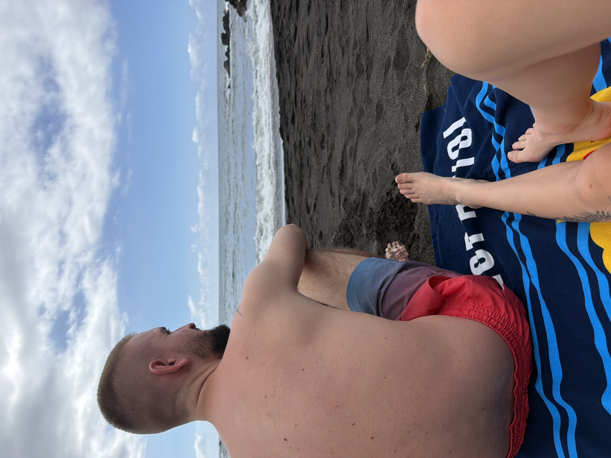 In Puerto de la Cruz, Spain, a man with a buzz cut and beard sits on a black sand beach, facing the ocean. He wears red and blue swim trunks. Beside him, on a blue towel with yellow and blue stripes, rest a woman's bare legs and feet. The ocean is visible in the background, with waves gently rolling towards the shore. The sky is partly cloudy with a mix of blue and white.