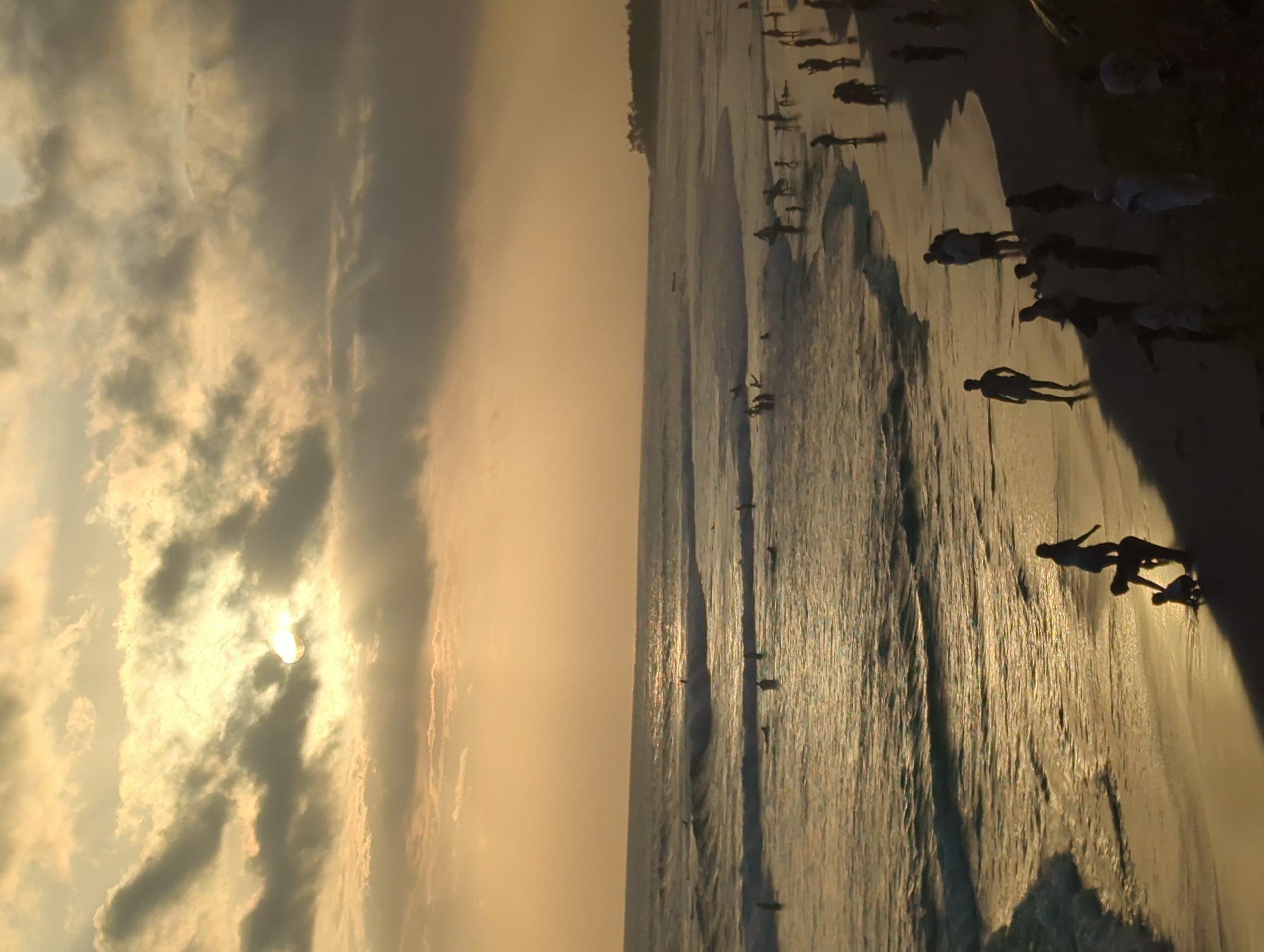 The image depicts a beach scene, likely at sunset, in North Kuta, Indonesia. The sky is filled with clouds, partially obscuring the sun, casting a golden hue over the scene. The beach is populated with numerous silhouetted figures, some appearing to be walking or standing near the shoreline. The waves are visible, reflecting the sunlight, and indicate an ocean setting. The sand appears damp and reflective. The presence of multiple people suggests a public beach area.
