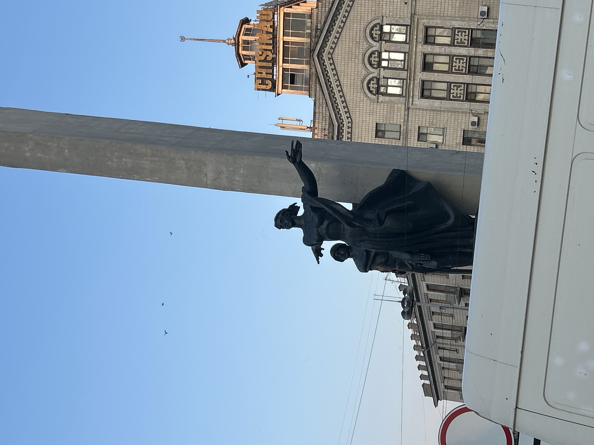 The image captures a statue of two figures, a woman and a man, against a clear blue sky with a tall concrete obelisk to their right. The woman, in the foreground, is depicted with an outstretched arm, pointing. Behind her, the man is partially visible. In the background, a building with the word "CHISINAU" prominently displayed on its upper facade stands out. The architecture of the building suggests a post-Soviet era style. The lower portion of the image is occupied by the white roof of a vehicle, partially obscuring the base of the statue and a red and white circular traffic sign. Several power lines are visible crisscrossing the sky. A few birds are scattered across the sky.