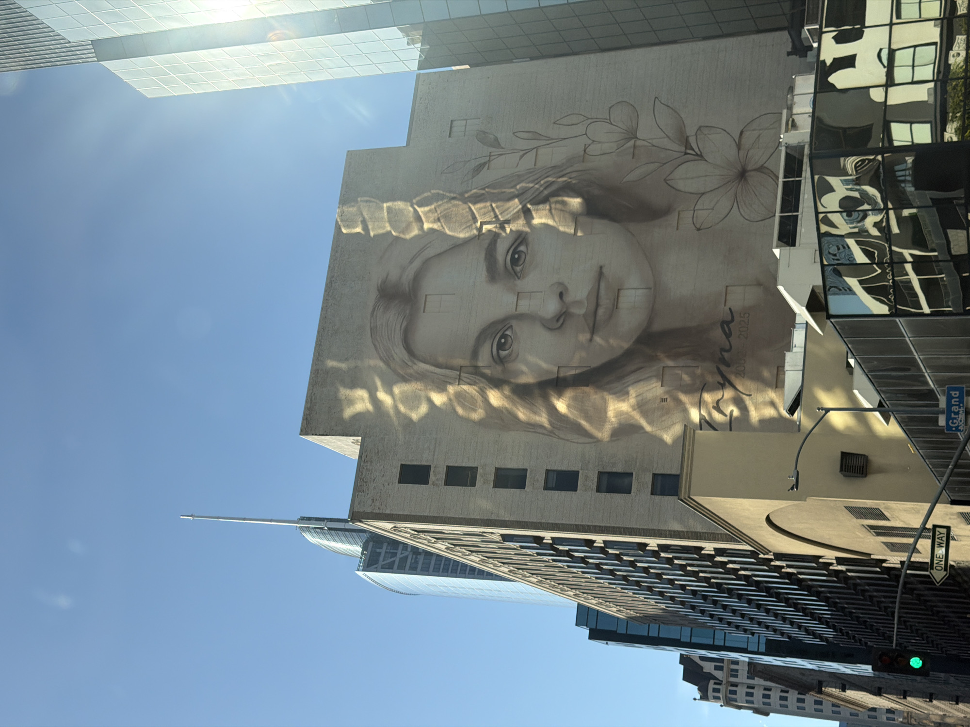 The image depicts a street scene in Los Angeles, California. Visible are several buildings, including a taller building with a mural of a woman's face painted on its side. The mural features a portrait of a girl or young woman with foliage around her face. Other buildings with glass and steel facades are visible, reflecting the sky. Street signs are visible, including a 'ONE WAY' sign and a street name sign that partially reads 'Grand'. A traffic light is lit in green. The sky is clear and blue, with a hint of sunshine at the top of the frame.