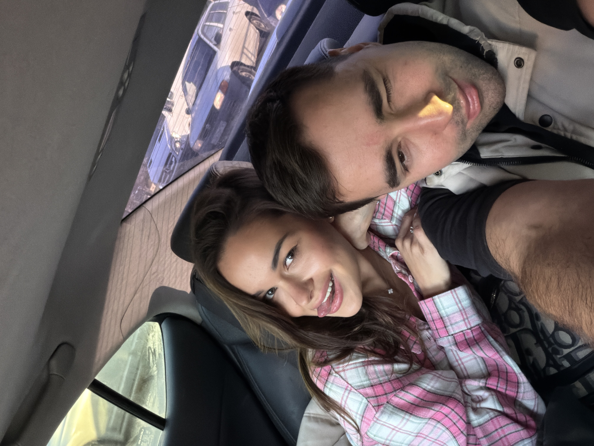 A man and a woman are in what appears to be the back seat of a car. The woman is wearing a pink and white checkered shirt and smiling while sticking her tongue out. The man is wearing a light-colored jacket and has his eyes partially closed. Through the car window, there are other cars, possibly indicating they are in transit. It is impossible to determine if the location is Krasnoyarsk, Russia from this image alone.