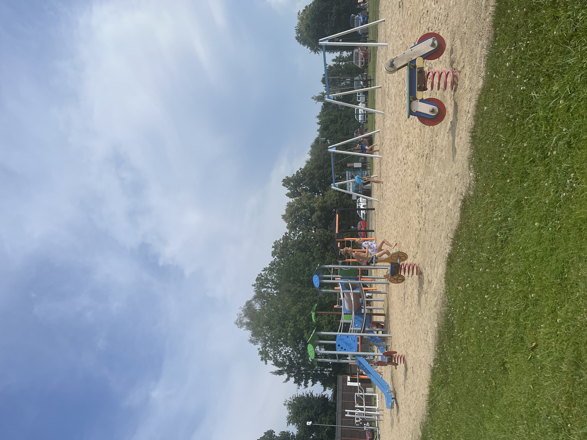 A wide-angle shot captures a vibrant playground on a bright, sunny day. The scene is dominated by a sandy ground cover, with a lush green grassy area on the right. Several playground structures are visible, including swings, a slide, and spring riders. In the foreground, a red and blue spring rider with a yellow seat is partially visible. Further back, a complex blue and orange structure with a slide and climbing elements stands prominently.
Two young children are actively engaged in play. One child, wearing a blue top and shorts, is swinging. Another child, in a patterned swimsuit, is seated on a yellow spring rider, appearing to be in motion. The presence of children suggests a playful and lively atmosphere.
The background features a line of mature green trees, indicating a potentially wooded or park-like setting. Beyond the trees, several cars are parked, and some buildings are visible, hinting at a community or recreational area. The sky above is a clear blue, with scattered white clouds, suggesting a pleasant daytime weather condition.
There is visible text on the blue structure, which appears to read "Fiyglen.pl" and "Fiyglen.pl" again. The overall impression is one of a joyful summer day at a well-equipped playground.