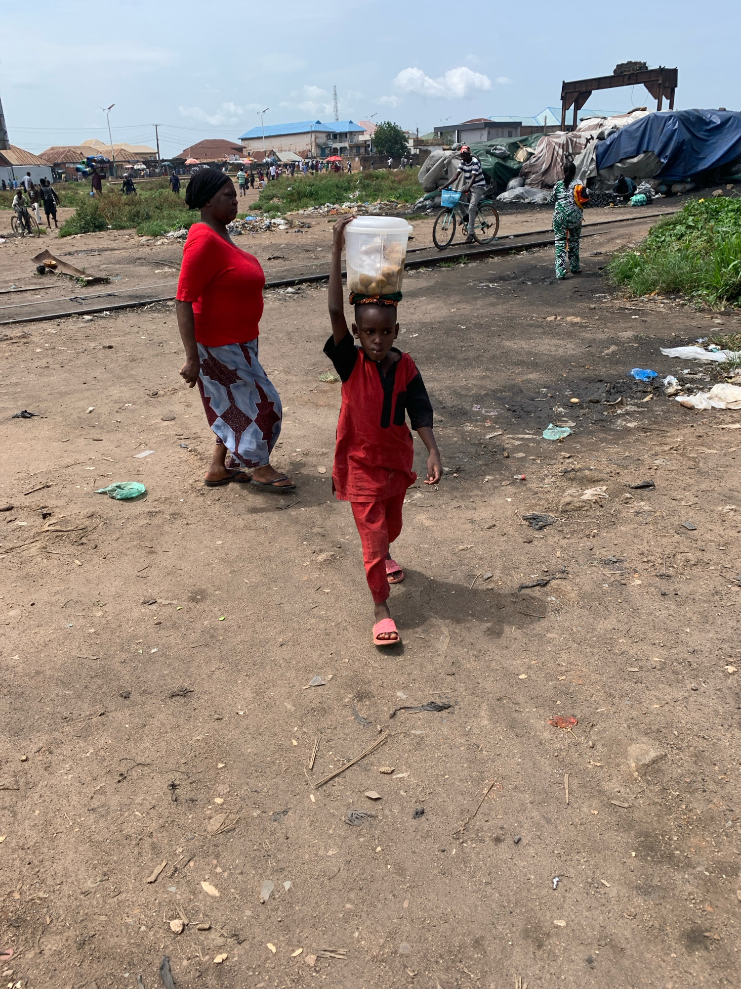In Bukuru, Nigeria, a young child, possibly a boy, walks towards the camera, carrying a clear plastic container filled with what appears to be food items on their head. The child is wearing a red tunic with black trim and matching red pants, along with pink flip-flops. To the left of the child, an adult woman, dressed in a red shirt and a patterned skirt, walks away from the camera, looking back over her shoulder. She wears a headscarf. The environment is an unpaved area, likely a street or open lot, with dirt, scattered debris, and some vegetation visible. In the background, several buildings, including structures with blue and red roofs, are present, suggesting a populated urban or semi-urban setting. A bicycle is also visible in the mid-ground, with a person riding it. Several large, blue tarpaulins are draped over unseen objects in the background, possibly indicating storage or construction. The scene appears to be outdoors under daylight conditions with a cloudy sky.