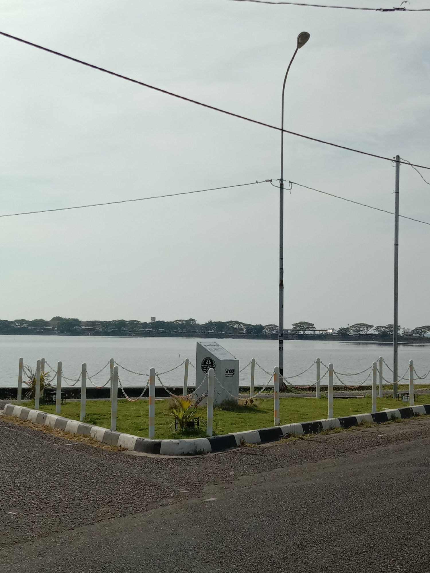 The image captures a serene outdoor scene by a body of water in Lhokseumawe, Indonesia, likely a lake or large river. The foreground features an asphalt road with a checkered black and white curb, bordering a small, triangular patch of green grass. This grassy area is enclosed by white posts connected by chains, serving as a decorative barrier. Two small, leafy plants are visible within the grassy patch.
Dominating the middle ground, immediately behind the barrier, is a white rectangular monument or sign. On its front face, the word "Sengaja" is clearly visible in a modern sans-serif font. Below this, there's a circular emblem or logo, which appears to incorporate symbols like a tree or plant and possibly a boat or wave design. Other text below "Sengaja" is present but too blurry to decipher accurately, though "Senyaman" and "Sejahtera" might be partially readable.
Beyond the immediate foreground and monument, the expanse of the calm, greyish-blue water stretches across the image. The far bank is lined with a dense growth of trees and some distant buildings, suggesting a populated area. Overhead, two tall utility poles stand, one on the left with a curved street lamp, and another on the right. Multiple power lines or communication cables crisscross the upper portion of the sky, which is bright and largely clear but appears slightly hazy or overcast.
The time of day appears to be daytime, likely late morning or early afternoon, characterized by bright, diffused sunlight. There are no people visible, and no specific activity is taking place, creating a peaceful and quiet atmosphere.