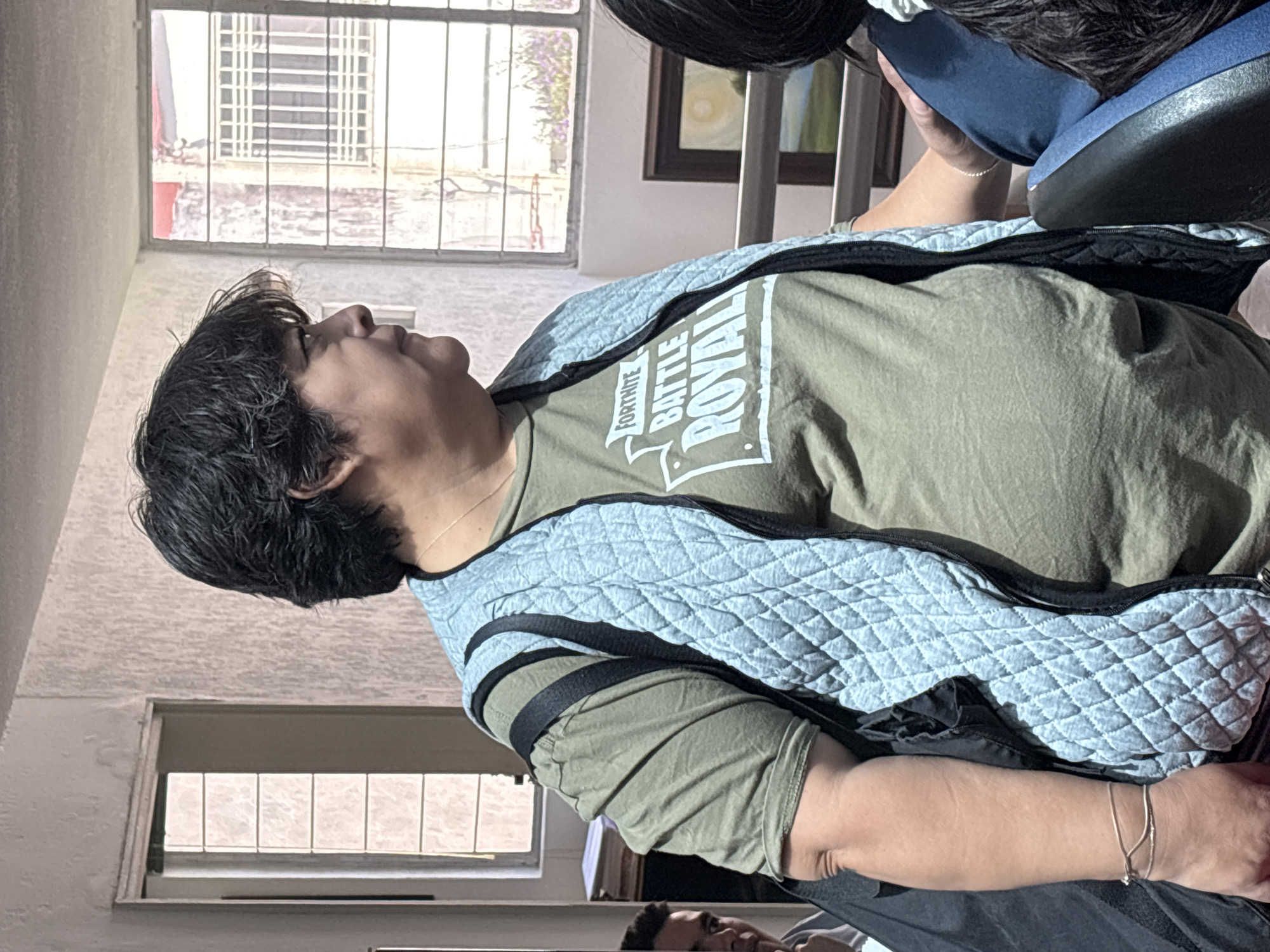 The image, taken indoors in Puebla, Mexico, features a young person with dark, medium-length hair, seen in profile looking upwards. They are wearing an olive green t-shirt with the white text "FORTNITE BATTLE ROYAL" prominently displayed, layered under a light blue quilted vest with black trim. A thin silver chain is visible around their neck, and a delicate silver bracelet adorns their right wrist. Their expression appears calm or contemplative as they gaze upwards.
The setting is a brightly lit room, likely during the daytime, with natural light streaming through large windows in the background. These windows are fitted with white metal grates, and beyond them, parts of other buildings and some distant greenery are visible. The walls are light-colored, and a framed picture can be seen on the wall in the upper background. A blue and black headrest or chair back is partially visible at the top right, suggesting the person is leaning against it. Another person's head is partially visible at the bottom of the frame, also seemingly looking upwards. The overall scene conveys a casual and relaxed atmosphere.