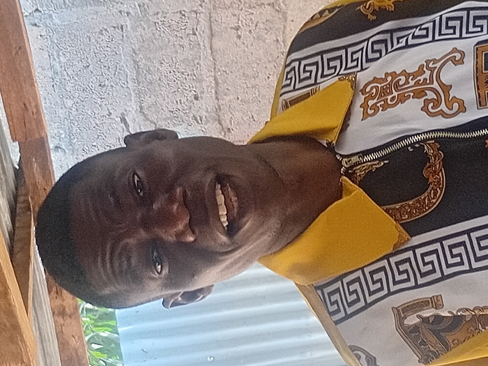 A frontal portrait shot depicts an adult male in Ayangba, Nigeria. The subject has short, dark hair and dark eyes, looking directly at the camera. His mouth is slightly open, revealing several teeth. He wears a collared, long-sleeved shirt with a central metallic zipper. The shirt features a solid yellow collar and upper shoulder sections. The main body of the shirt displays a pattern of alternating vertical panels: white panels with black Greek key borders, and black panels containing intricate gold and brown baroque-style scrollwork and a distinct gold chain motif. The background consists of a light-colored, textured concrete or brick wall on the right, a corrugated metal roof supported by visible wooden beams overhead, and a hint of green foliage on the far left. The subject is stationary.