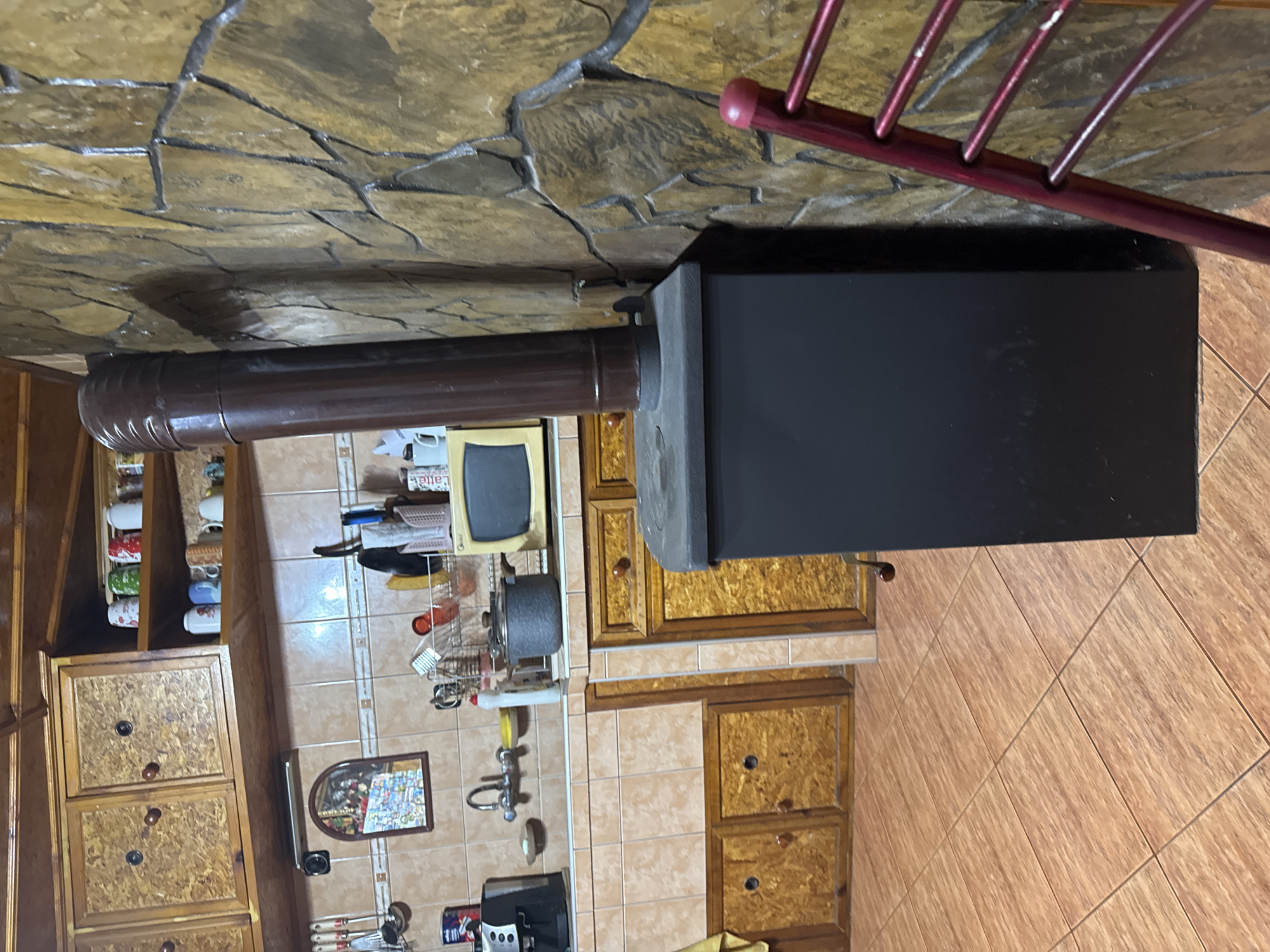 This image captures the interior of a domestic kitchen located in Sarnevo, Bulgaria. The scene is dominated by a black wood-burning stove situated against a textured stone wall on the right side of the frame. A dark brown stovepipe extends vertically from the stove and connects to the stone-clad wall, which features irregular, earth-toned stones and dark grout lines.
To the left of the stove, a kitchen area is visible, comprising a tiled countertop and backsplash. The backsplash consists of light brown rectangular tiles, some with a subtle pattern, and a decorative border tile line. A chrome-plated faucet is installed above a visible sink basin. A rack holding various kitchen utensils hangs on the tiled wall to the left of the sink. Above the sink, a small, arched mirror with a decorative frame reflects an indistinct colorful pattern.
On the countertop, a black coffee or espresso machine is positioned on the far left. Towards the center-right of the countertop, a grey cooking pot rests, and further right, a compact, beige microwave oven is visible. Above the countertop, wooden upper cabinets with doors made of oriented strand board (OSB) or a similar composite wood material with round wooden knobs are mounted. Adjacent to these cabinets, open wooden shelving holds an assortment of colorful mugs and cups.
The ceiling features exposed wooden beams and planks. The floor is covered with light brown, rectangular ceramic tiles laid in a staggered pattern. In the lower right foreground, a portion of a red metal object, possibly a chair or a drying rack, is partially visible. No individuals are present in the image.
