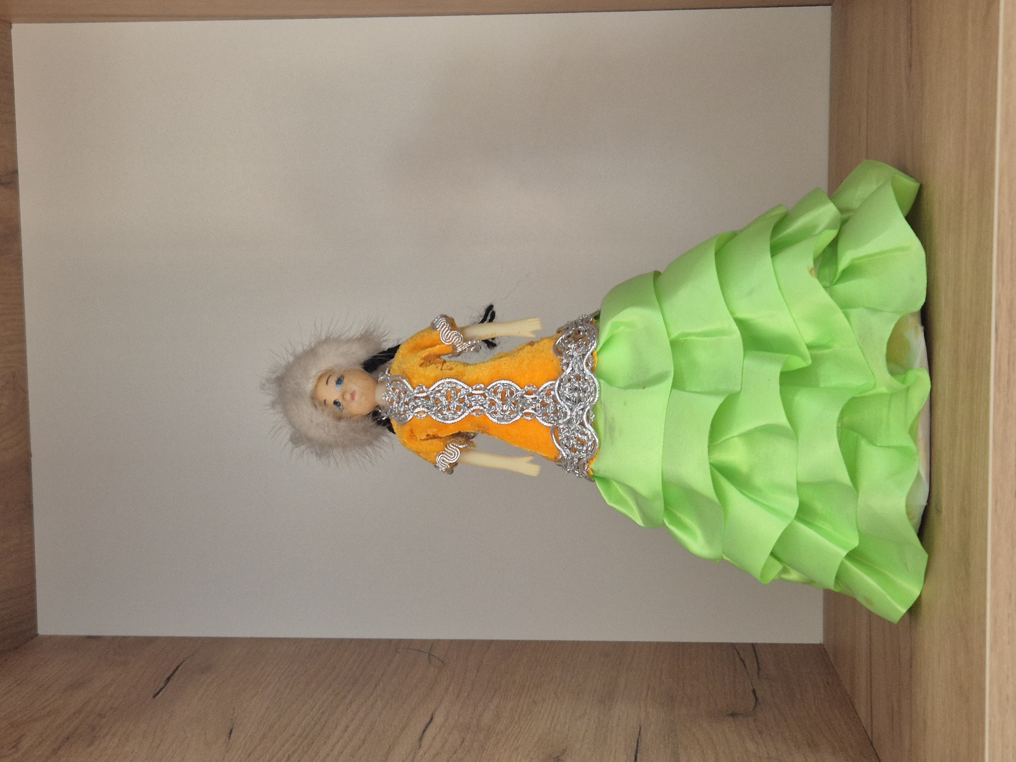A doll dressed in traditional clothing stands on a wooden shelf. The doll has blonde hair and blue eyes and wears a furry white hat. Her dress is orange with silver trim and a green, ruffled skirt. The background is a plain, light-colored wall. The shelf is situated within a cabinet with wooden sides. There are no people or activities depicted in the image. The scene is static and appears to be an indoor display. The location in Kokshetau, Kazakhstan, is not discernible from the image itself.