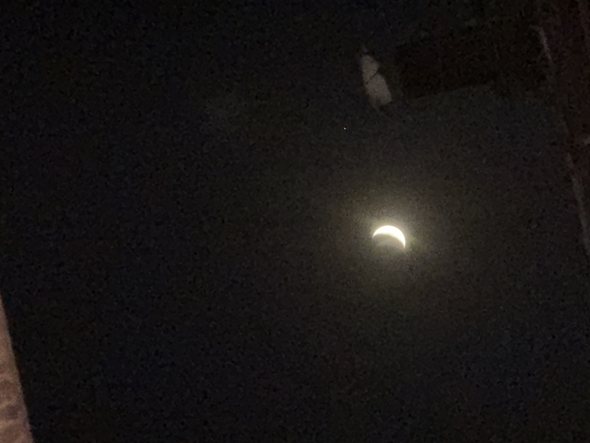 The image shows a crescent moon in a dark sky. The moon is the brightest object in the frame, with a clear sliver of light visible. There is a faint, distant light to the right of the moon, possibly a star or another celestial body. The sky is a deep black, with some subtle texture suggesting it's not entirely clear. The bottom portion of the image appears to show a blurry, dark structure, possibly a rooftop or building, with some lighter areas indicating it might be reflecting light. The overall impression is of a nighttime sky, with the moon as the central focus. No specific landmarks or features are identifiable that would confirm its location in Yerevan, Armenia.