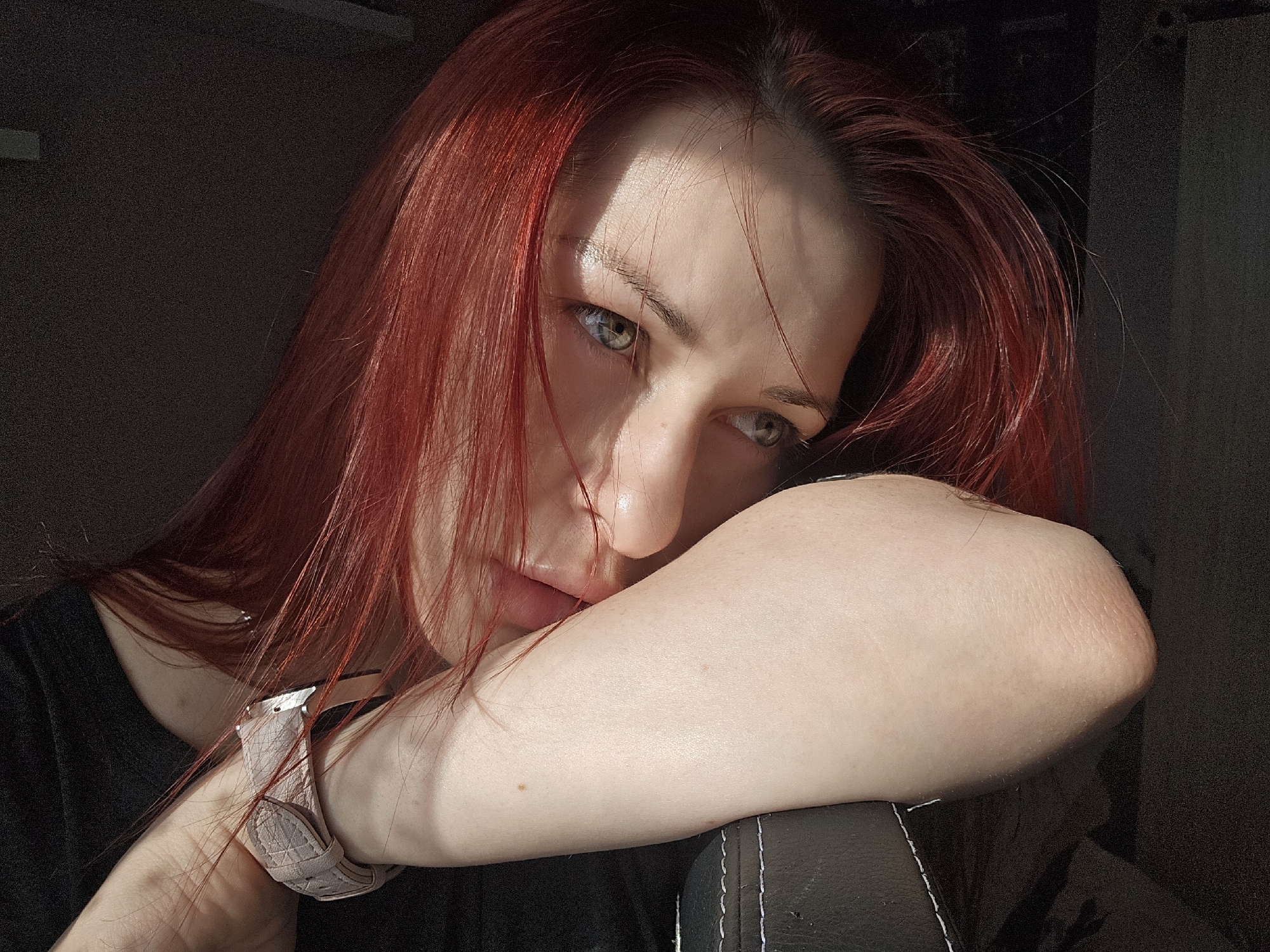 A fair-skinned woman with auburn hair rests her head on her arm. She has light eyes and is wearing a dark shirt. A watch with a light-colored strap is on her wrist. Her arm rests on a dark, possibly leather, surface. The lighting is uneven, with shadows and bright areas, suggesting indoor light. The image provides no information about the location, Burgas, Bulgaria.