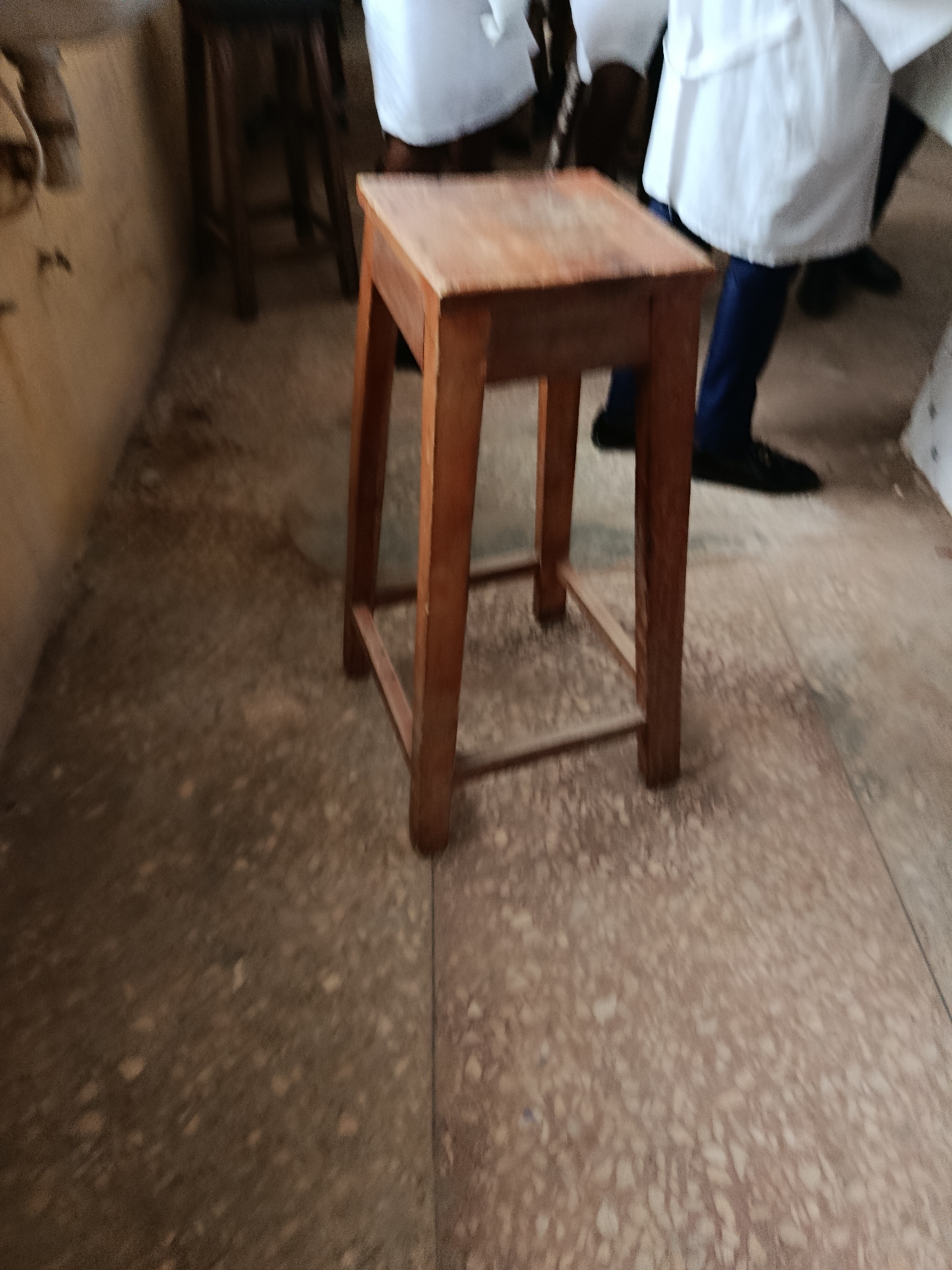 The image shows a wooden stool in the foreground, with its legs and seat clearly visible. The floor is a speckled, light-colored material, possibly terrazzo or polished concrete. In the background, parts of people wearing white lab coats and dark pants are visible, suggesting a laboratory or medical setting. The lighting is somewhat dim, casting shadows and creating a muted atmosphere. The overall impression is of an indoor space, possibly a classroom or research facility. The specific location in Ukpor, Nigeria, is not indicated by any visual cues within the image itself.