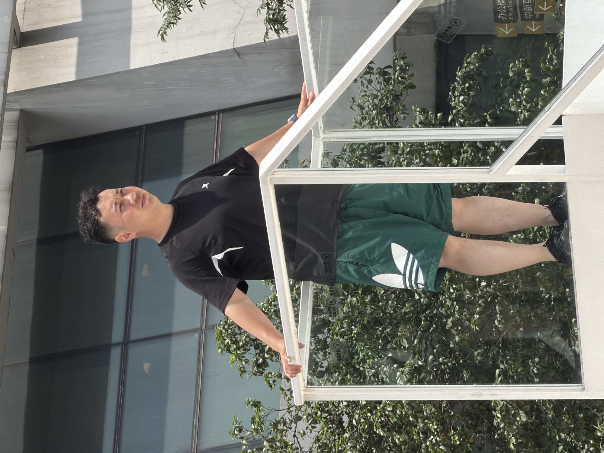 A light-skinned man with short, dark, curly hair stands on a raised white platform. He is wearing a black t-shirt with a white emblem on the left chest and green athletic shorts with a large white Adidas logo on the left leg. He is also wearing black athletic shoes. The man is looking to his left, with his left hand resting on a clear glass railing. In the background, there are green leafy trees and a modern building with large windows. Signs in Chinese are visible on the glass railing, indicating "Office Tower" and directions to "Middle China". The scene appears to be outdoors in a city environment, possibly Yuzhong District, China, given the signage. The lighting suggests it is daytime.