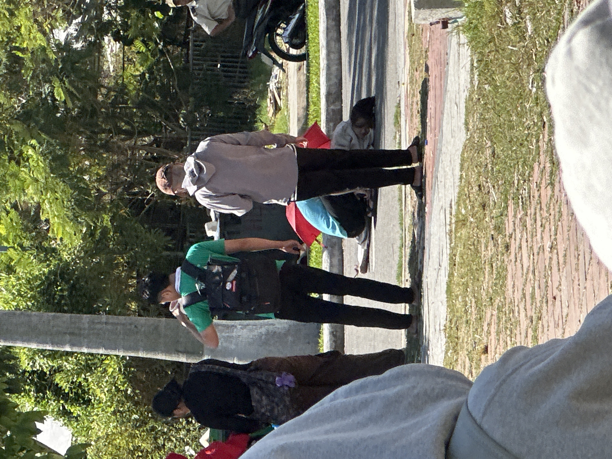 In the city of Đà Nẵng, Vietnam, on a sunny day, several individuals are visible. A person in a green t-shirt with a backpack stands near a large tree, appearing to drink from a cup. To their left, another person in black clothing stands facing away from the camera, a patterned bag with a purple stuffed toy hanging from it is visible. In the middle ground, two people are crouching near a red umbrella, with one person lying on the ground, seemingly injured. Further back, a person in a light-colored hooded sweatshirt stands, and to the right, a person in a light-colored uniform is standing near a motorcycle. The scene takes place outdoors with vegetation and paved surfaces visible.