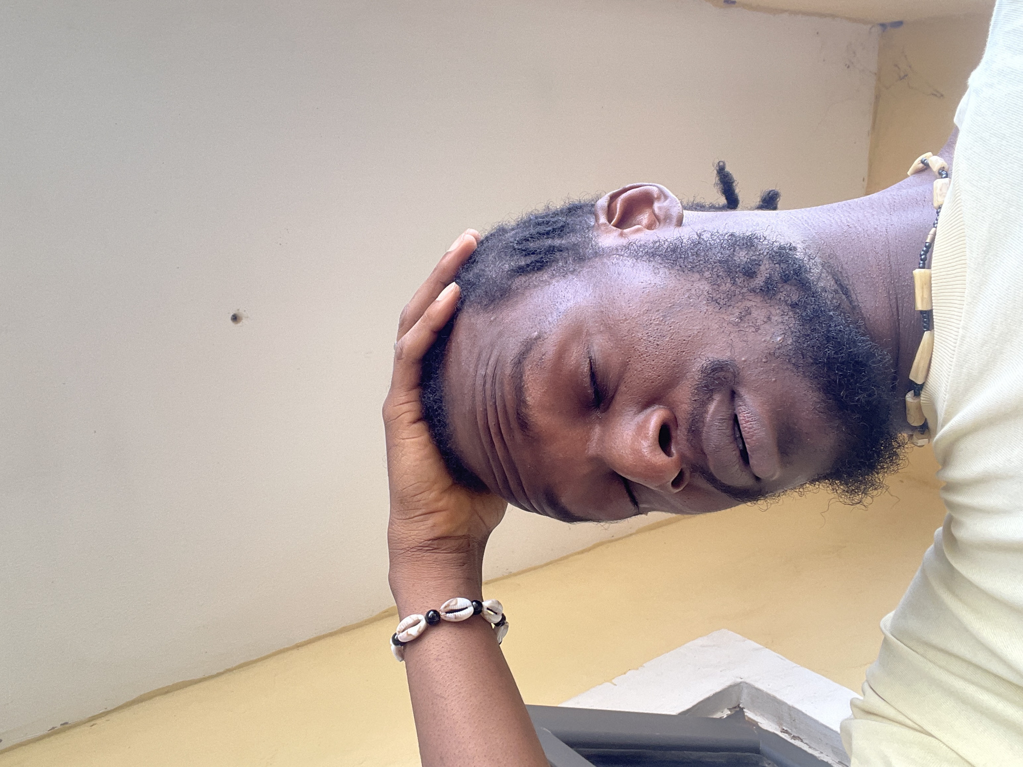 The image shows a close-up, upside-down view of a Black person's face and upper body. The person has closed eyes, a slight smile, and dreadlocks. They are wearing a light-colored shirt and a beaded necklace made of wood and black beads. On their wrist, they are wearing a bracelet made of cowrie shells and black beads. The background is a light-colored wall with a faint shadow, and a yellow surface below, suggesting an indoor or covered outdoor setting. The lighting appears natural, possibly from daylight. The person's hand is resting on their head, and their expression suggests a sense of peace or relaxation. No visible text is present in the image. The scene is likely in Abuja, Nigeria, as indicated by the provided location context.