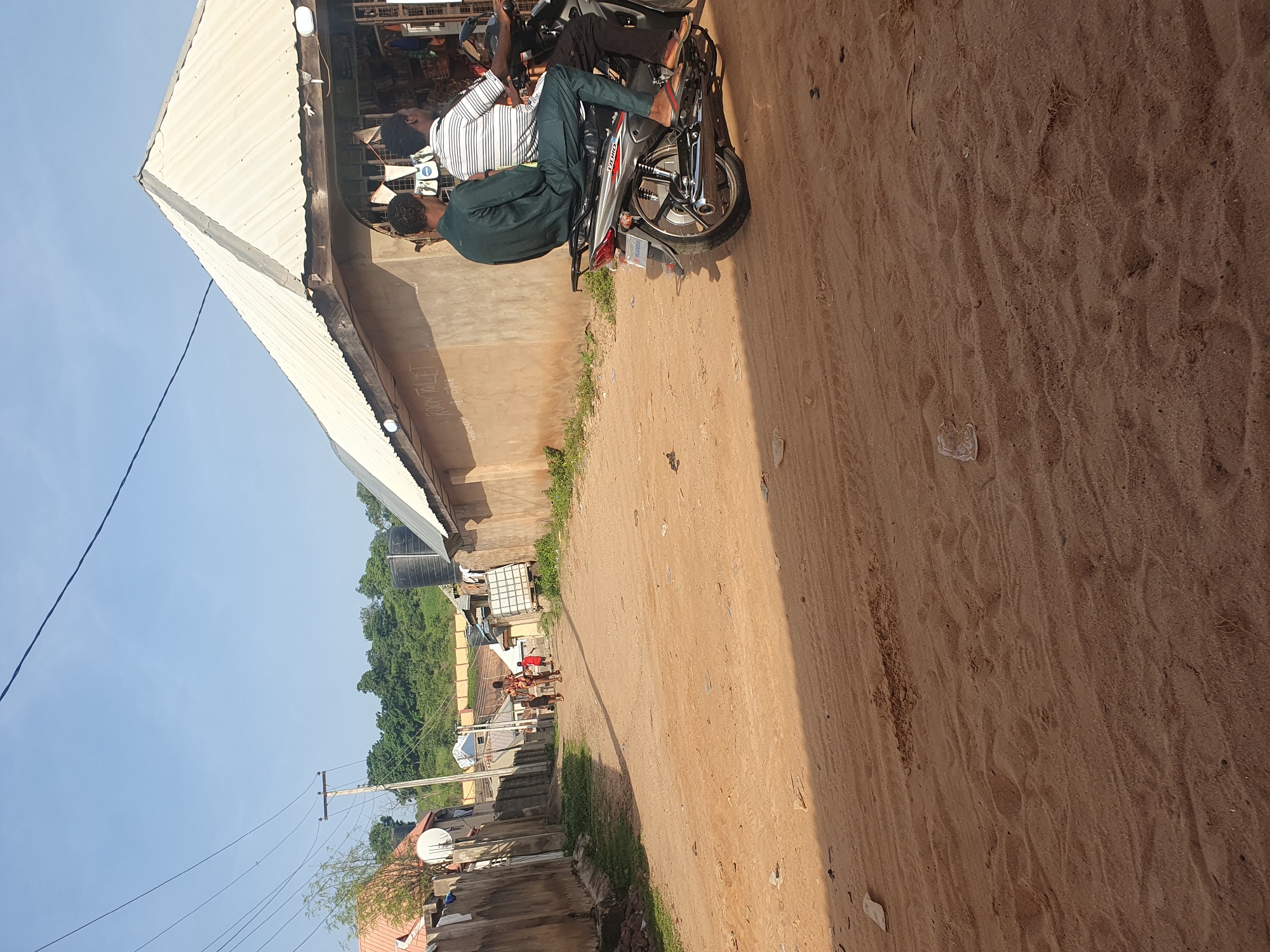 In Lokoja, Nigeria, a dirt road winds through a settlement. The road is unpaved and dusty, with tire tracks visible. On the right side of the road, a building with a corrugated metal roof and open storefronts is situated. Two men are riding a motorcycle in front of this building, with the driver in front and a passenger behind. The passenger is wearing a dark green long-sleeved shirt and trousers, while the driver is wearing a white and striped shirt and dark trousers. To the left of the road, a concrete wall runs alongside the street, and beyond that, more buildings and vegetation are visible. Further down the road, a group of people, including children, are walking away from the camera. The sky above is clear and blue, with power lines stretching across it. The overall scene depicts a typical street view in a Nigerian town, characterized by its unpaved roads, local businesses, and everyday activities.