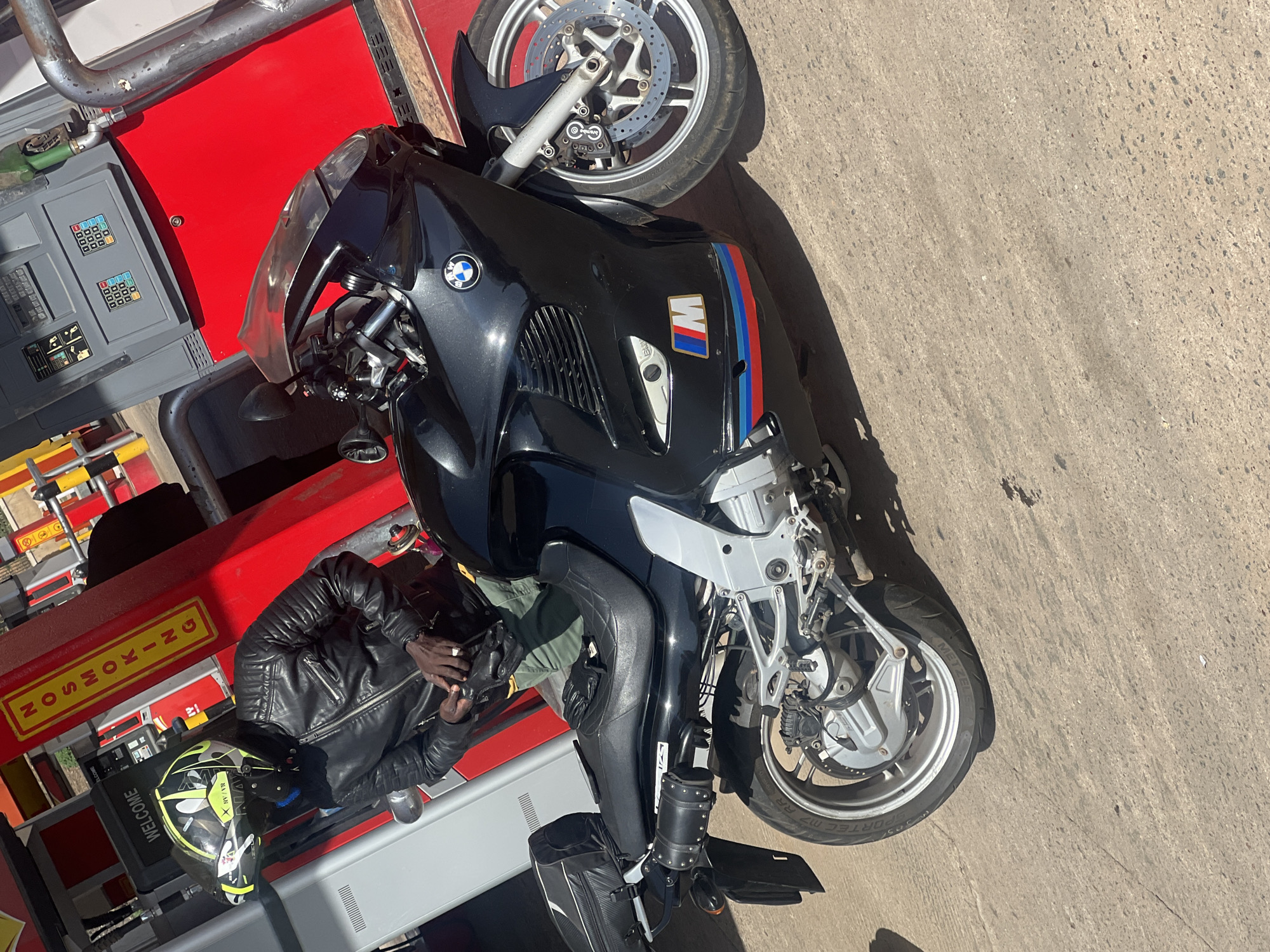The image shows a black BMW motorcycle parked next to a red and gray gas pump on a paved surface. A person wearing a black leather jacket and riding gloves is visible near the gas pump, alongside a helmet with green and yellow accents. The motorcycle features the BMW logo and the "M" badge with blue, red, and purple stripes, indicating the M performance model. The pump has a "NO SMOKING" sign in yellow and "WELCOME" written on it. The location is possibly in Gura Top, Nigeria based on the question prompt, though there are no specific identifiers in the image to confirm this.