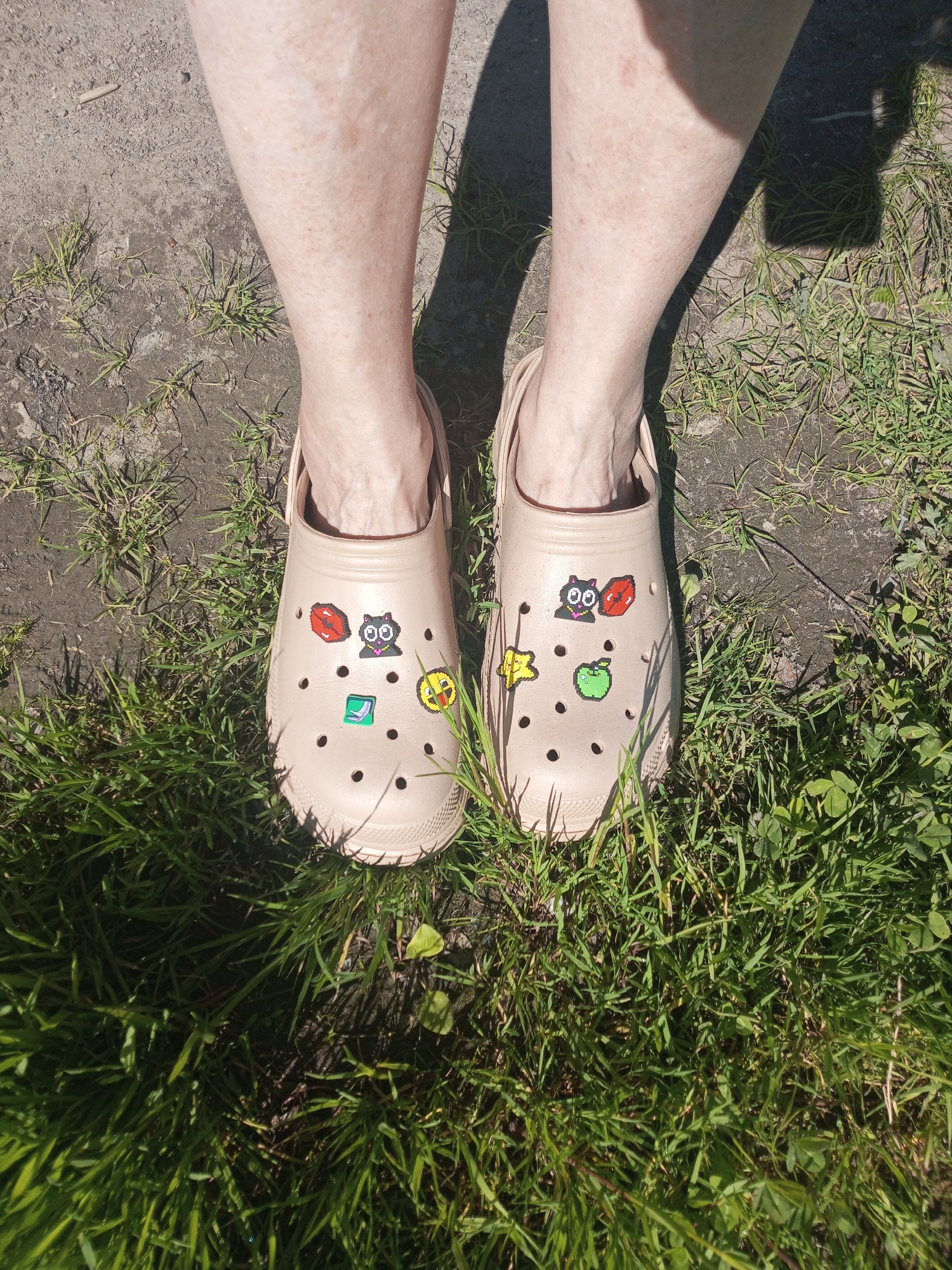 The image provides a top-down view of a person's lower legs and feet, clad in light beige Crocs clogs, standing on a patch of ground. The setting is outdoors, characterized by sparse green grass interspersed with patches of dry dirt or concrete.
Bright, direct sunlight is evident from the distinct, dark shadows cast by the legs and footwear onto the ground, indicating a clear and sunny day, likely during midday or early afternoon. The person's bare legs, visible from mid-calf down to the ankles, display fair skin with some visible freckles or minor blemishes.
The beige Crocs are customized with several colorful, pixelated Jibbitz charms. The left clog features a red pixelated mouth, a black pixelated cat with green eyes and a pink nose, a green pixelated square icon (resembling a message or paper symbol), and a yellow pixelated smiley face with a sticking-out tongue. The right clog is adorned with a black pixelated cat with green eyes, a red pixelated mouth, a yellow pixelated star, and a green pixelated apple.
The scene captures a static moment, showcasing the footwear and the immediate outdoor environment in Kommunar, Russia. No specific activity or emotion is discernible, and there is no visible text within the image.