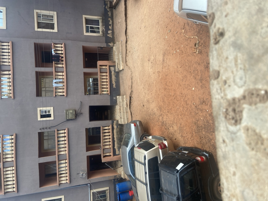 This image depicts a residential compound in Nsukka, Nigeria, viewed from an elevated perspective. The central subject is a three-story building with a gray plastered facade.
The building features multiple balconies on its first and second floors, each equipped with ornate, light-colored concrete balustrades. Rectangular windows with white frames are visible across all floors. Ground-level entrances, some fortified with black metal security gates, lead into the building. A utility meter box is mounted on the first-floor wall, positioned between two balconies. Light-colored trousers and dark garments are observed hanging to dry over the balustrades of two first-floor balconies.
The foreground and courtyard consist of unpaved, reddish-orange laterite soil, characteristic of the region, which appears uneven and somewhat barren. Three vehicles are parked consecutively on the left side of the unpaved area: a silver sedan, followed by a beige SUV, and a black SUV, all facing left. The rear section of a fourth silver vehicle is partially visible in the lower right corner of the frame. Near the left side of the building, two blue plastic drums and one red bucket are present on the ground. No individuals are visible within the scene. The image is taken during daylight hours.