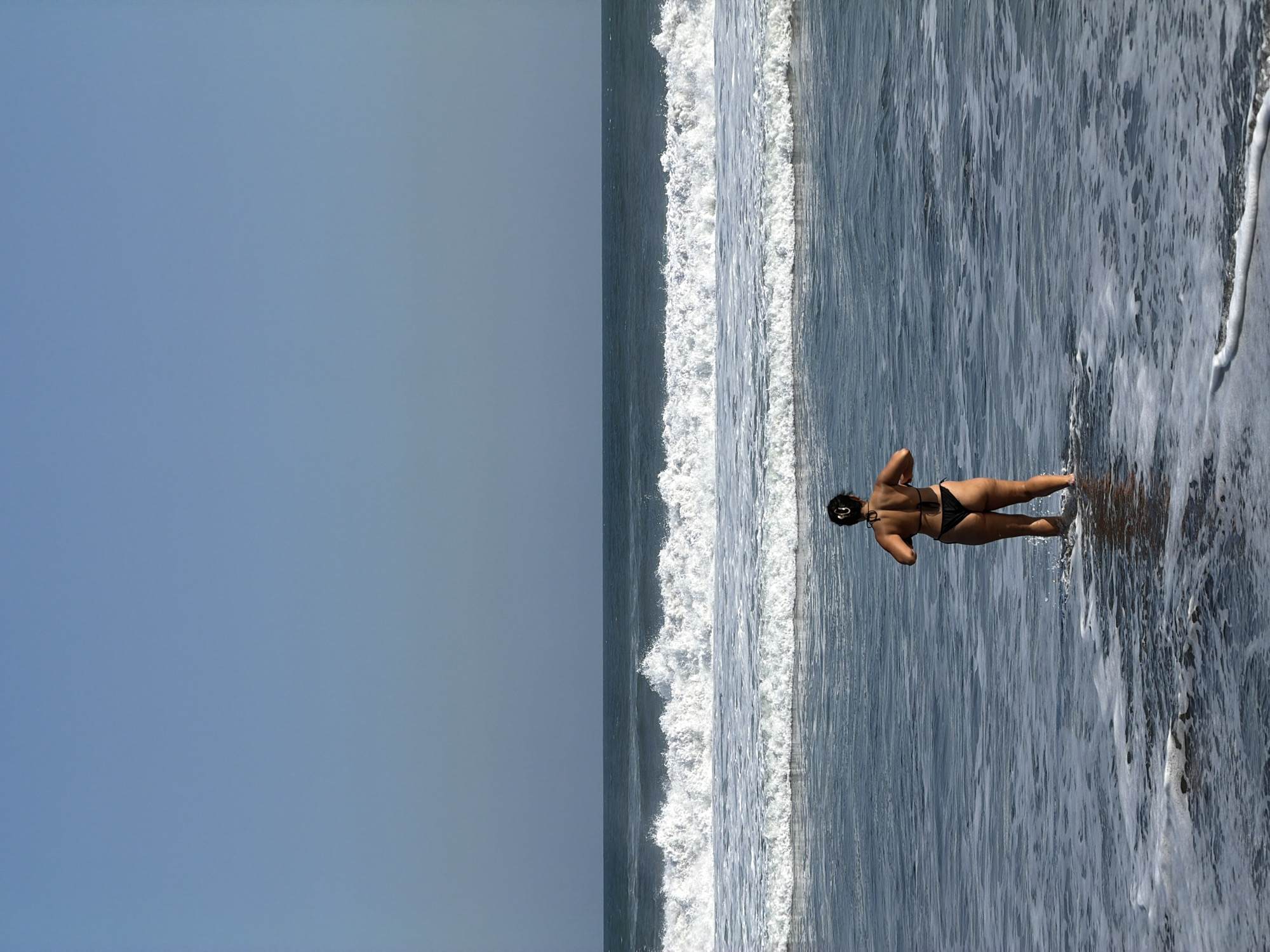 The image captures a serene ocean view in Selemadeg, Indonesia. An adult female, viewed from behind, stands in the shallow water with foam and ripples around her lower legs. She wears a black two-piece swimsuit. Her dark hair is pulled up into a bun at the back of her head. Her arms are bent at the elbows, with her hands positioned in front of her body as she looks out towards the sea. The sky above is a clear, uniform light blue, meeting a distinct horizon line that separates it from the ocean. In the mid-ground, multiple lines of white-capped waves break, with a prominent wave cresting across the scene. The water further out is a darker blue-grey and appears calmer.