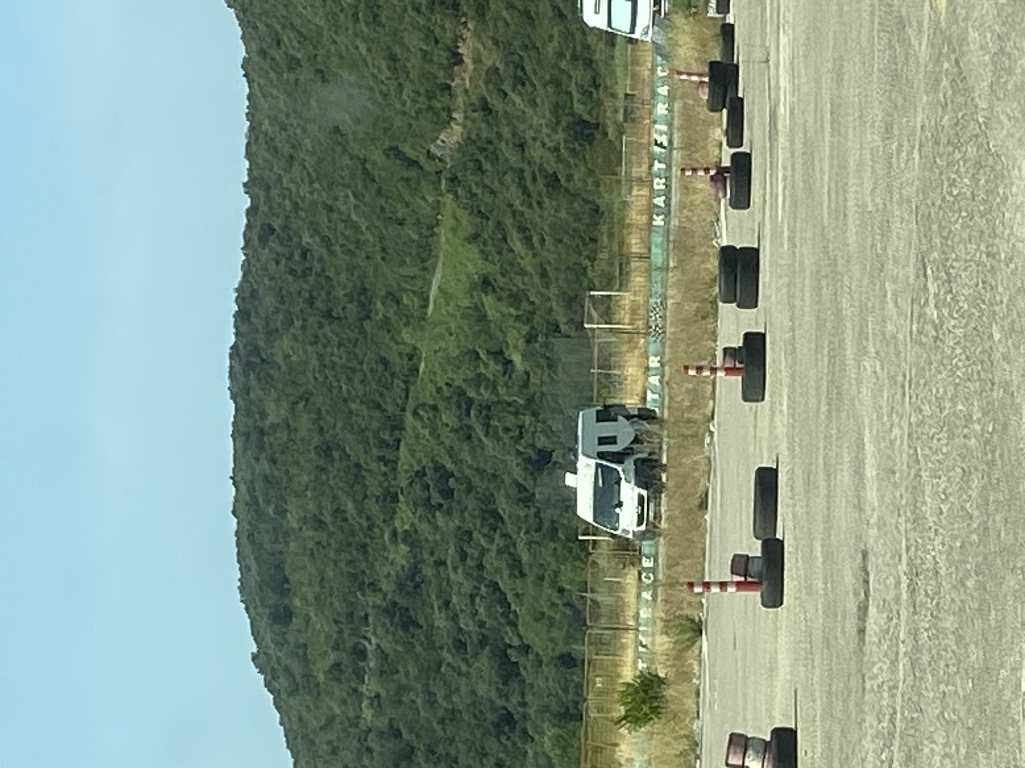 In Prijevor, Montenegro, a flat, paved area, likely a go-kart track, is depicted. The terrain is mostly flat with a large, tree-covered hill in the background. The sky is clear and blue. In the foreground, there are several stacks of tires, some with red and white striped cones placed on top. These tires and cones appear to be part of the track's safety or demarcation system. In the middle ground, a white van is parked. Behind the van, a fence runs horizontally across the image, with a checkered flag pattern visible on the lower section of the fence. The words "ACE" and "KART RACE" are visible on the fence. The overall scene suggests a functional but possibly quiet go-kart racing facility.