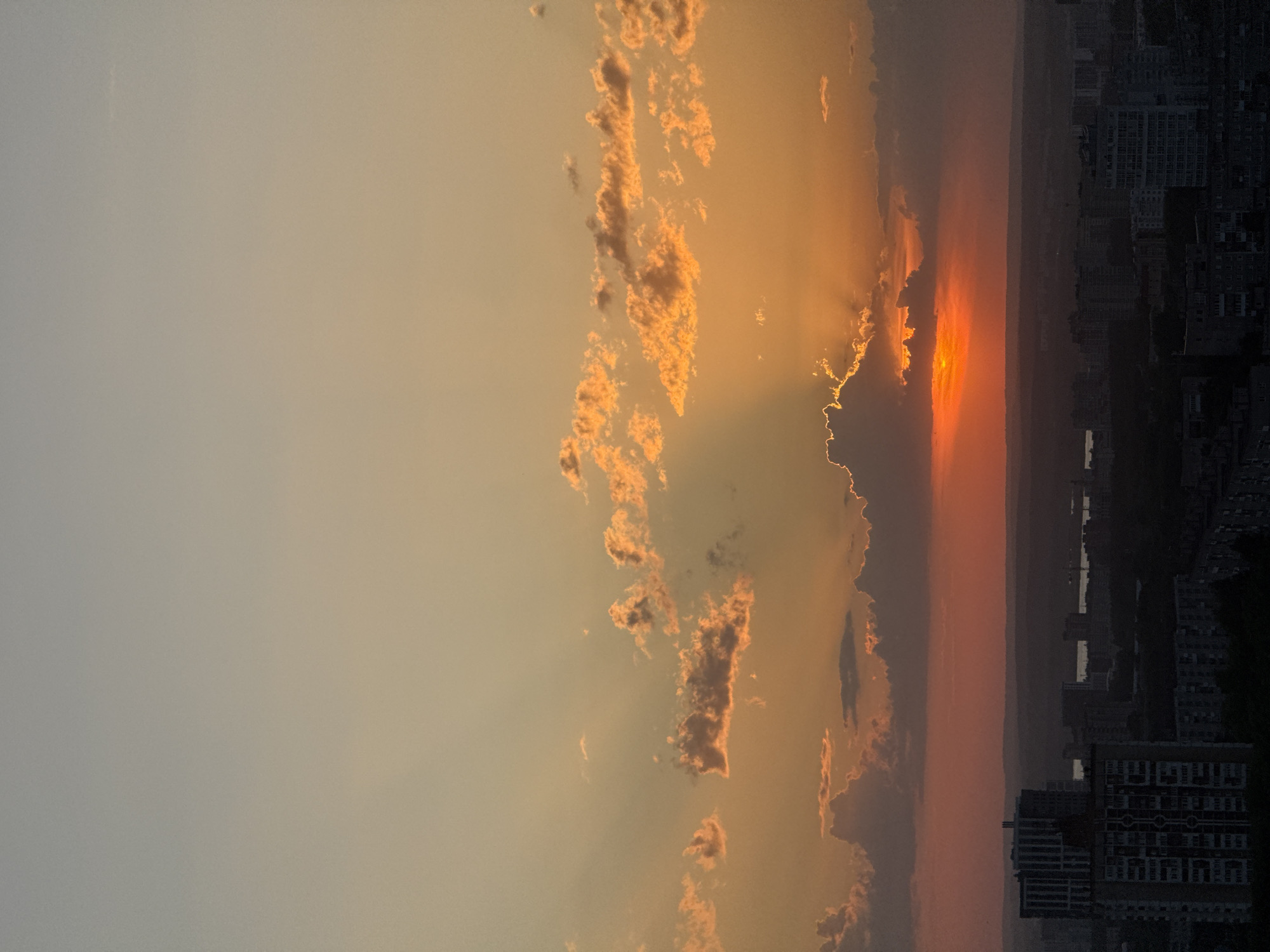 The image depicts a sunset over the city of Perm, Russia. The sky is filled with clouds, some of which are illuminated with orange and yellow light from the setting sun. The sun itself is visible through a break in the clouds, casting rays of light downward. In the lower right corner of the image, buildings of the city can be seen in silhouette. The river Kama can be seen in the distance, reflecting the colors of the sunset.