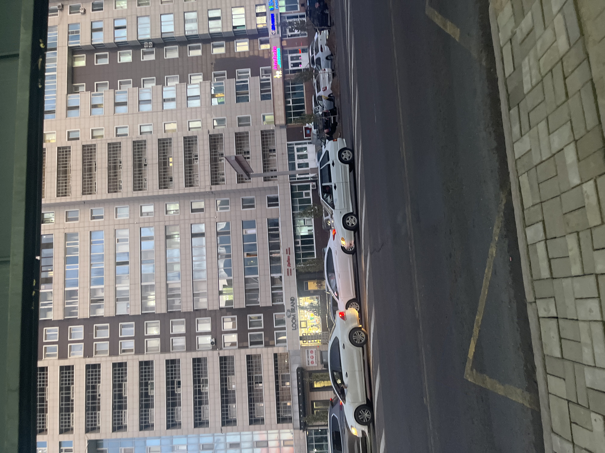 The image shows a street scene in front of a tall, modern apartment building. Cars are lined up on the street, some in traffic and some parked. The building has many windows and balconies, and some of the ground floor appears to be occupied by businesses. There is a sidewalk in the foreground made of brick pavers, and a yellow painted line on the asphalt street, indicating a no-parking zone. The sky is overcast and the lighting suggests it is either early morning or late afternoon. The overall scene depicts an urban environment.
