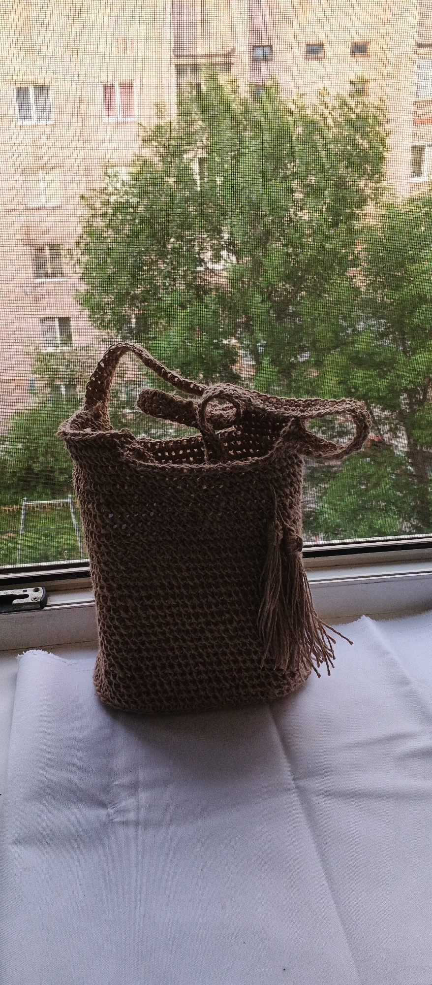 A close-up, slightly angled shot shows a crocheted, woven bag made of natural fibers. The bag is a neutral brown color, with a textured, open-weave pattern. It has two handles, one of which is a longer strap that crosses over the opening of the bag. A tassel made of the same fiber hangs from the right side of the bag. The bag is placed on a white cloth surface.
In the background, a window with a screen is visible. Through the screen, a brick building with several windows is apparent, some with curtains. Lush green trees fill the mid-ground, with their branches visible behind the building. A glimpse of a playground with a swing set can be seen to the left, behind the window frame. The scene is illuminated by natural light, suggesting it is daytime. The overall setting appears to be a domestic environment, possibly an apartment or house, with a view overlooking an urban or suburban area. This image was captured on July 15, 2024, at 4:37 PM in Izhevsk, Russia.