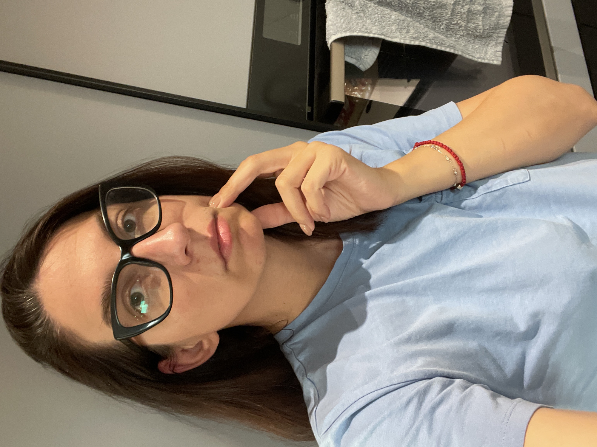 A woman with brown hair and glasses is in a room. She is wearing a light blue shirt and a red bracelet with silver charms. The woman is touching her cheek with her index finger. Behind her is a wall and a black oven. A gray towel is hanging near the oven. The location is in Sofia, Bulgaria, but this is not definitively discernible.