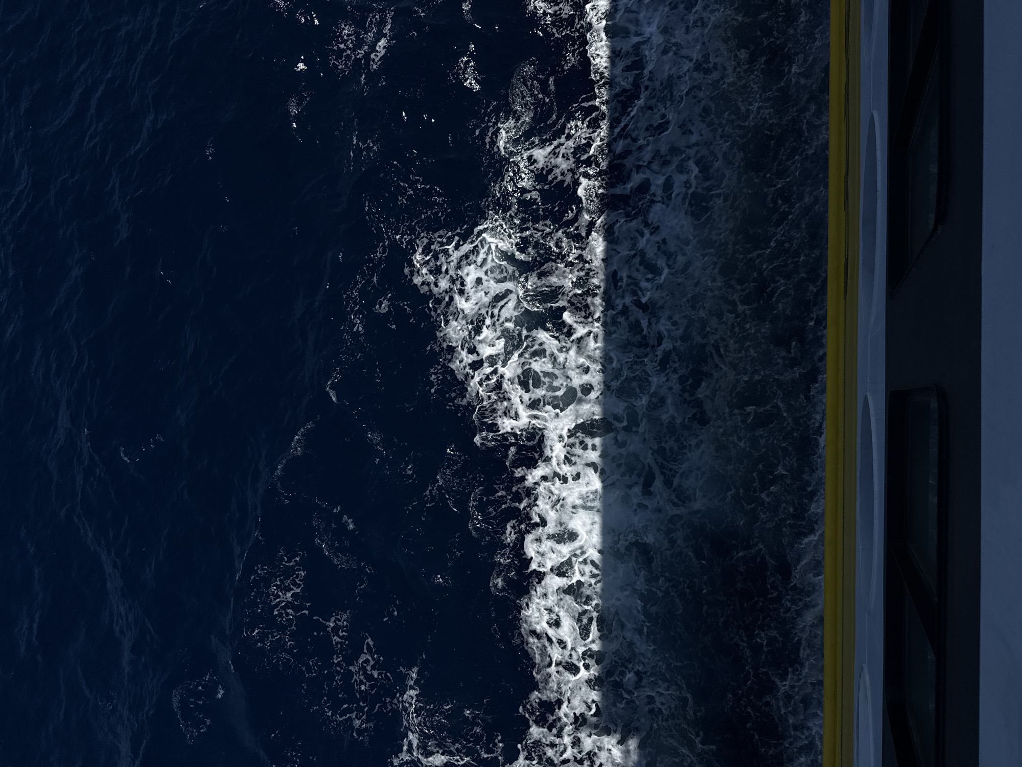 The image is an aerial, top-down view of dark blue ocean water with white foam, likely from a boat or ship's wake. The water transitions from choppy foam near the bottom of the frame to smoother, deeper blue water towards the top. At the very bottom of the frame, a yellow railing is visible, suggesting the vantage point is from the deck of a vessel. Below the railing, a dark surface with what appear to be rectangular windows or hatches is also visible. The scene appears to be set in the Municipal Unit of Kastro - Kyllini, Greece, though this specific location cannot be definitively confirmed from the visual information alone. There are no people or explicit actions depicted in the image.