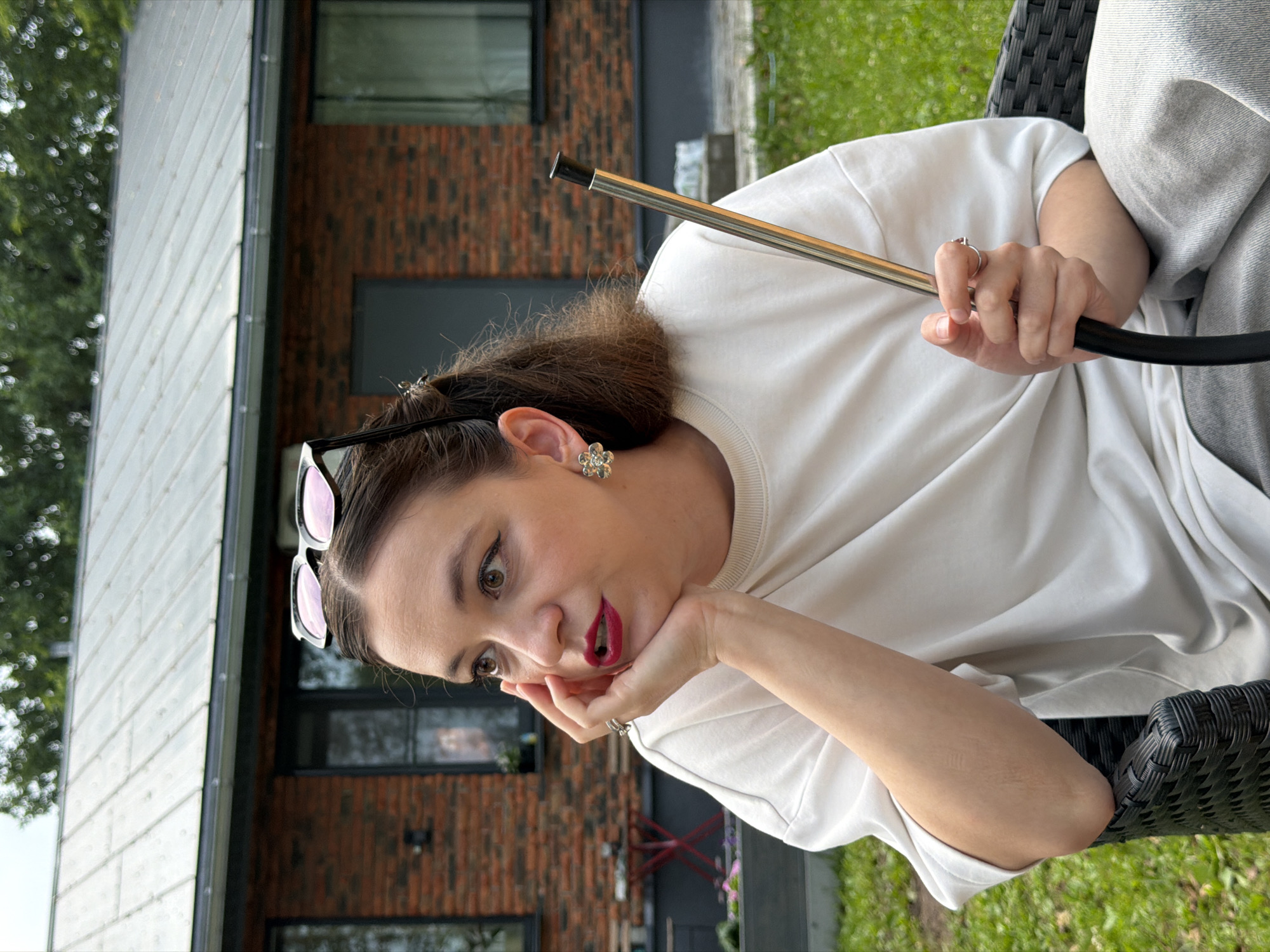 The media file shows a medium shot of a young woman sitting outdoors in a black woven chair. She is wearing a white t-shirt, light grey sweatpants, and sunglasses perched on her head. The woman has dark brown hair, is wearing flower-shaped earrings, and has bright red lipstick. She is leaning her head on her hand, with her elbow resting on her arm. Her expression appears curious or surprised. She is holding a long, metallic object, possibly a hookah hose.
The background features a modern brick building with large windows and a white-tiled roof, suggesting a residential or commercial structure. Green grass and trees are visible, indicating an outdoor setting during daylight hours, likely a warm day given the outdoor activity. The overall scene suggests a relaxed, informal setting. There is no visible text in the image. The provided location context is Chaikovsky, Russia.