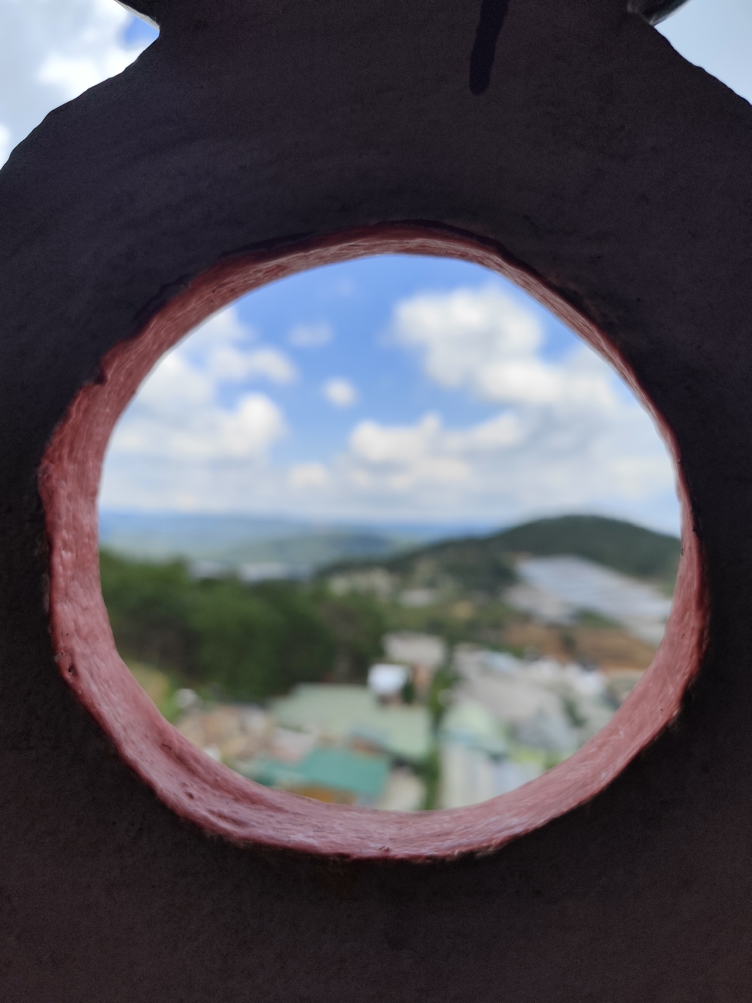 The image is a photograph taken through a circular opening in a dark, textured object, possibly a pot or a wall feature. The opening is lined with a rough, reddish-pink material. Through this opening, a blurred landscape is visible. The landscape appears to be a hillside with buildings and vegetation, set against a backdrop of a blue sky with white clouds. The location is identified as Lộc Quý, Vietnam. The focus is on the circular frame, with the distant scenery being out of focus, creating a depth-of-field effect. There are no people or animals visible in the image.