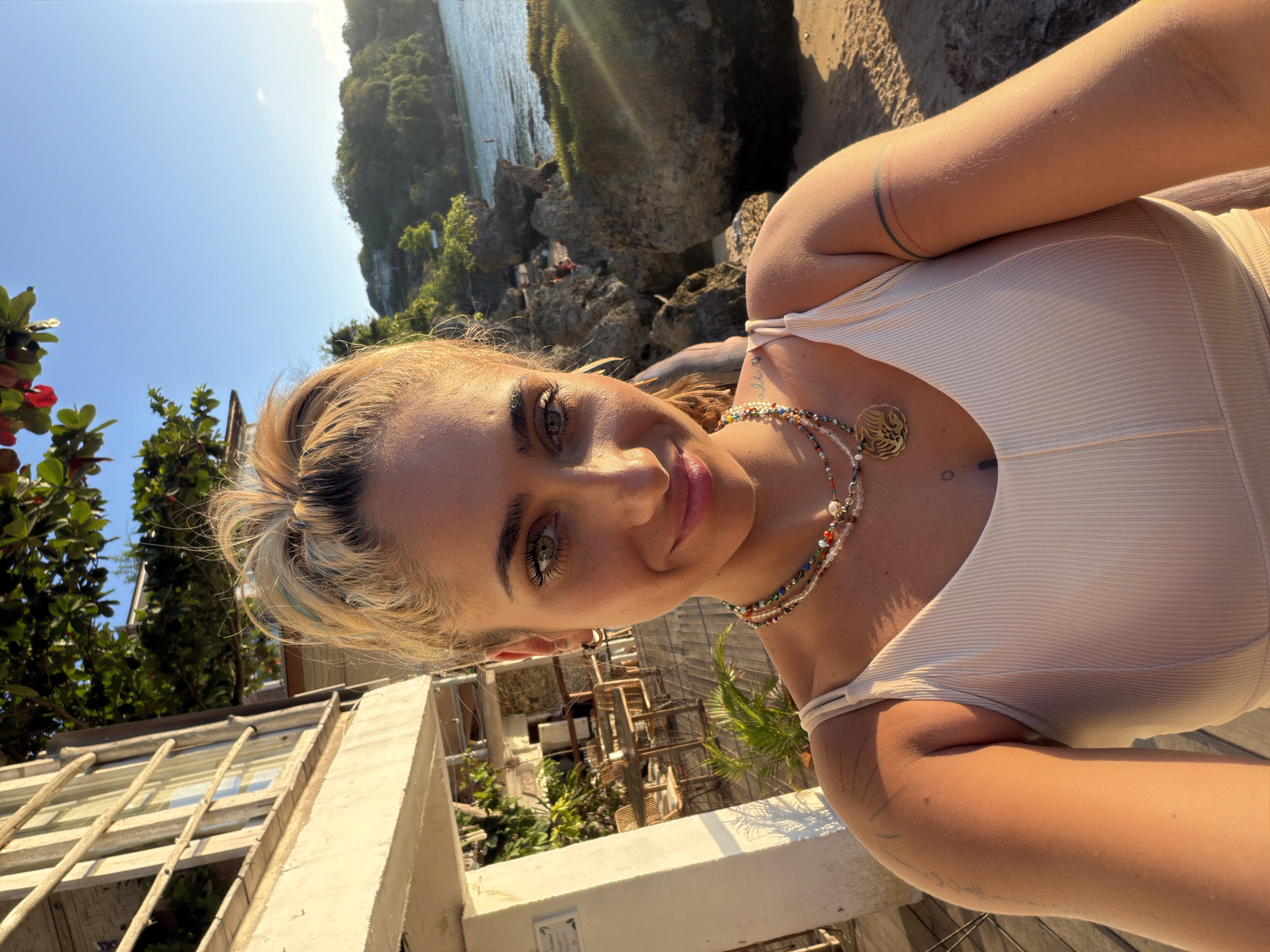 A woman with blonde hair pulled back in a ponytail, green eyes, and a smiling expression is taking a selfie. She is wearing a light-colored tank top and layered necklaces. She has tattoos on her arms. The background shows a beach scene with rocks, the ocean, and lush green foliage. To the left, there is a building with white pillars and wooden railings, with tables and chairs visible on a deck. The image appears to have been taken in Kuta Selatan, Indonesia, with the setting sun casting a warm glow on the scene.