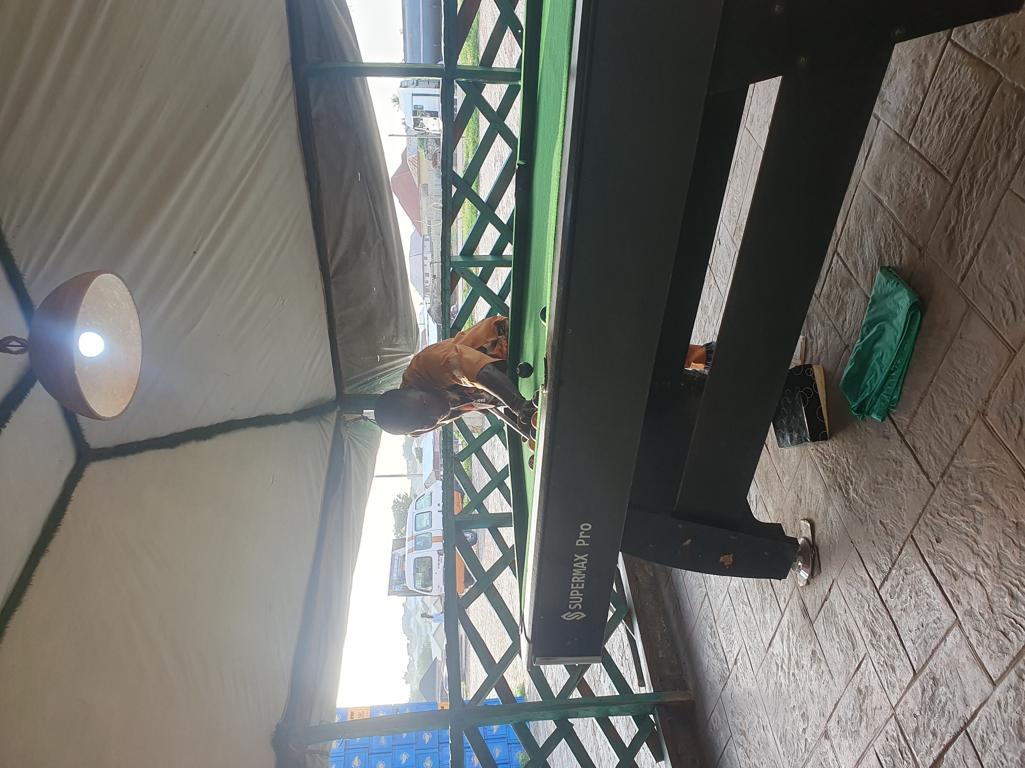 A man in an orange shirt plays billiards under a white tent at an outdoor establishment in Lokoja, Nigeria. The pool table, marked "SUPERMAX Pro," is green and appears well-used. The man is in the process of taking a shot. The tent is supported by dark poles and features a light fixture overhead. The ground is paved with textured tiles. In the background, a wooden fence separates the area from a road with vehicles, buildings, and vegetation. A green folded cloth is visible on the ground near the pool table.