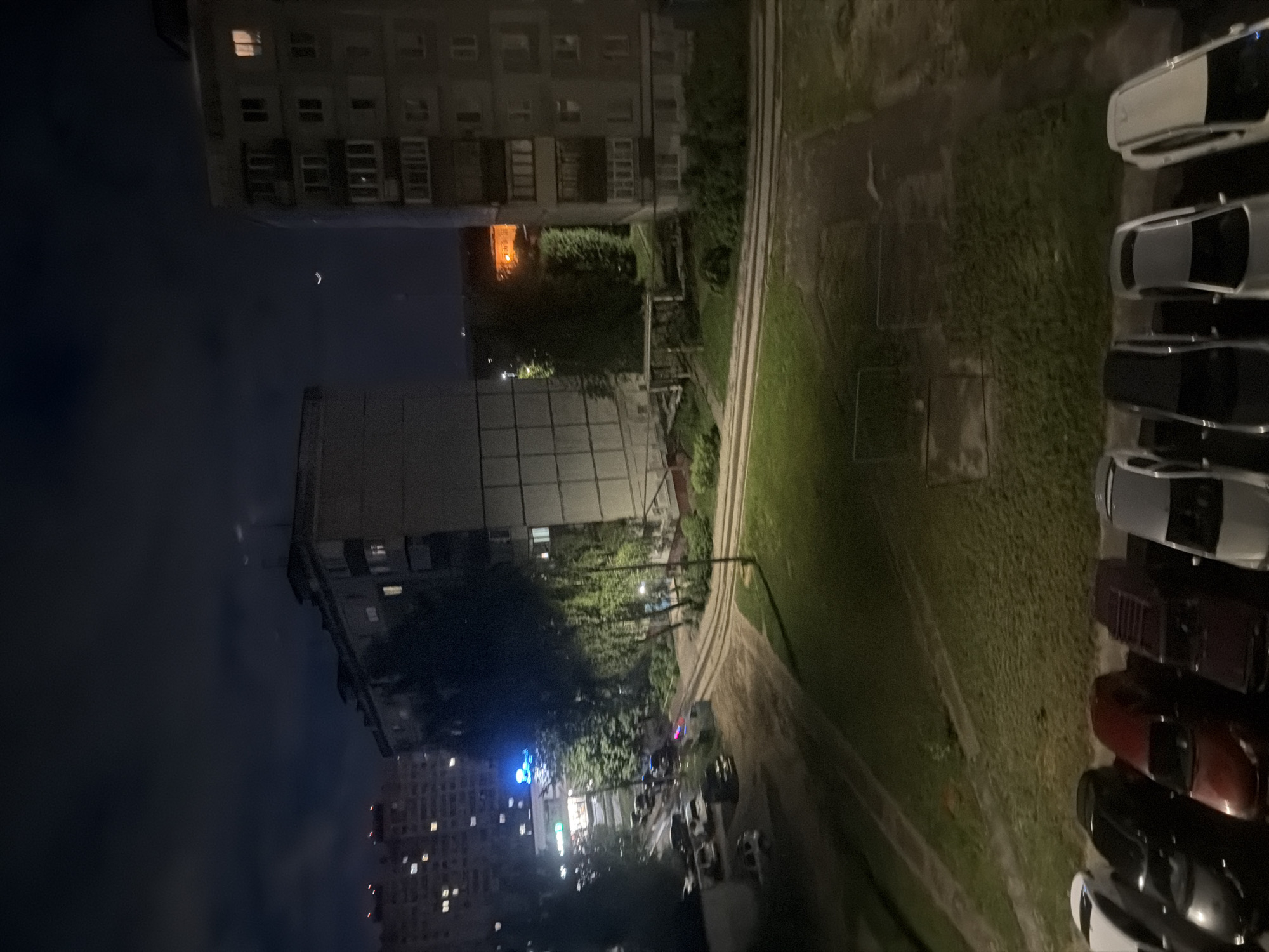 This media file depicts an outdoor scene at night, likely in a residential area of Nizhny Novgorod, Russia. The primary subjects are residential buildings and a row of parked cars.
The scene is dominated by a dark sky, suggesting it is nighttime. The lighting is low, with some artificial lights emanating from the windows of the apartment buildings. One building, in the foreground and oriented vertically, appears to be a multi-story apartment block with visible windows, some of which are illuminated. Another taller apartment building is visible in the distance, also with illuminated windows. To the right of the buildings, there is a patch of grass and a paved area, where several cars are parked in a line. These cars are of various colors, including white, silver, dark red, and black. Some of the cars have their headlights or interior lights on.
Notable details include the presence of trees and some greenery between the buildings and the parking area, indicating a somewhat urban but not entirely concrete environment. The presence of apartment buildings suggests a residential setting. There is no visible text in the image. The overall mood is quiet and still, consistent with a nighttime scene in a residential area. The orientation of the image is also unusual, appearing to be taken from an elevated perspective and rotated.
