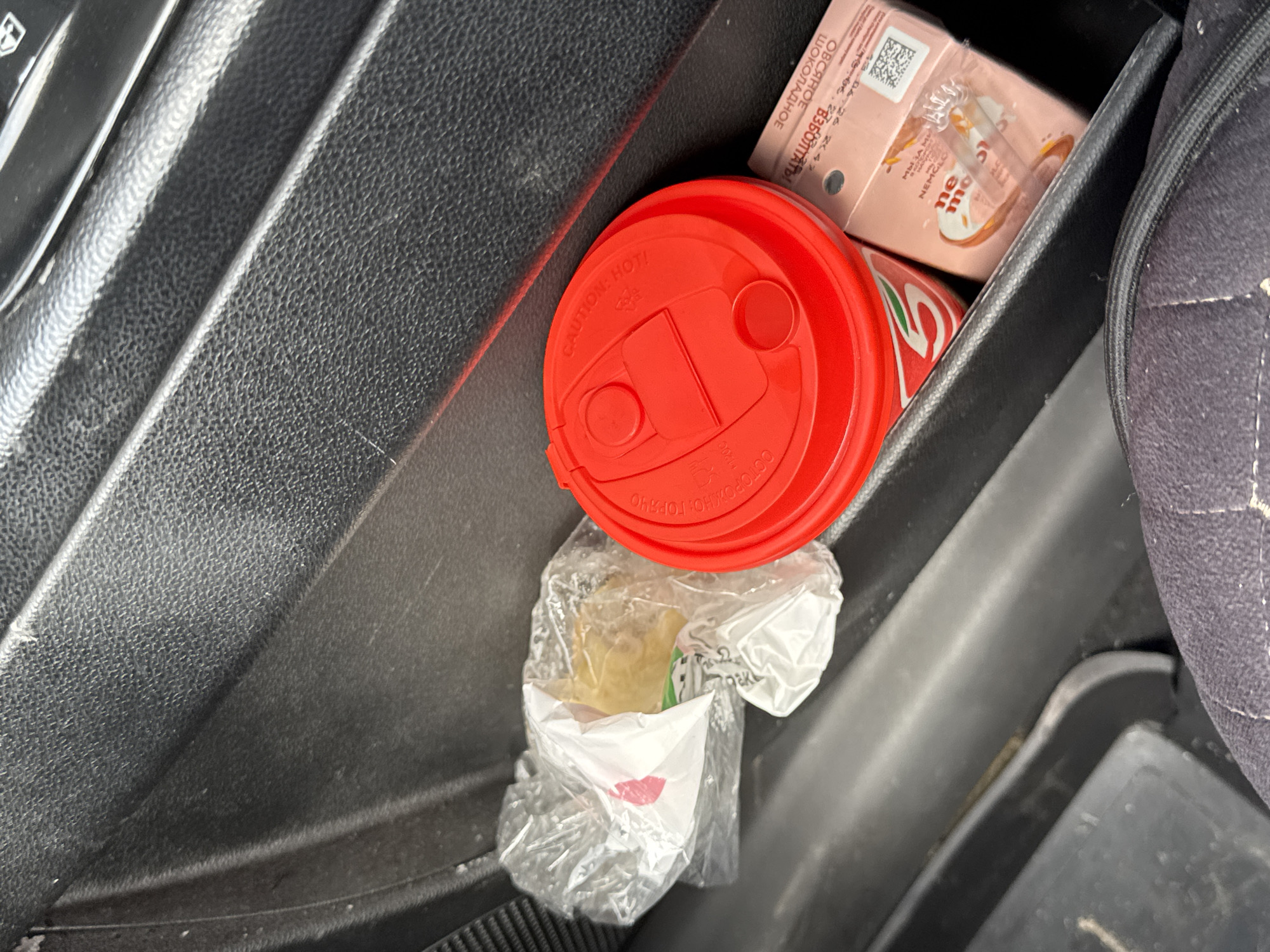 This is a close-up, eye-level, medium shot of a car's center console storage compartment in Ust'-Syny, Russia. The compartment is black and textured. Within the compartment, there are three items. On the left, a white and clear plastic bag contains a light brown, crumpled item, possibly food. To the right of the bag is a red paper cup with a red lid, appearing to be a drink container with a white "5" logo visible. To the right of the cup is a small, rectangular pink package with text and an image of a pastry or dessert on it. The overall scene is indoors, likely in a vehicle.