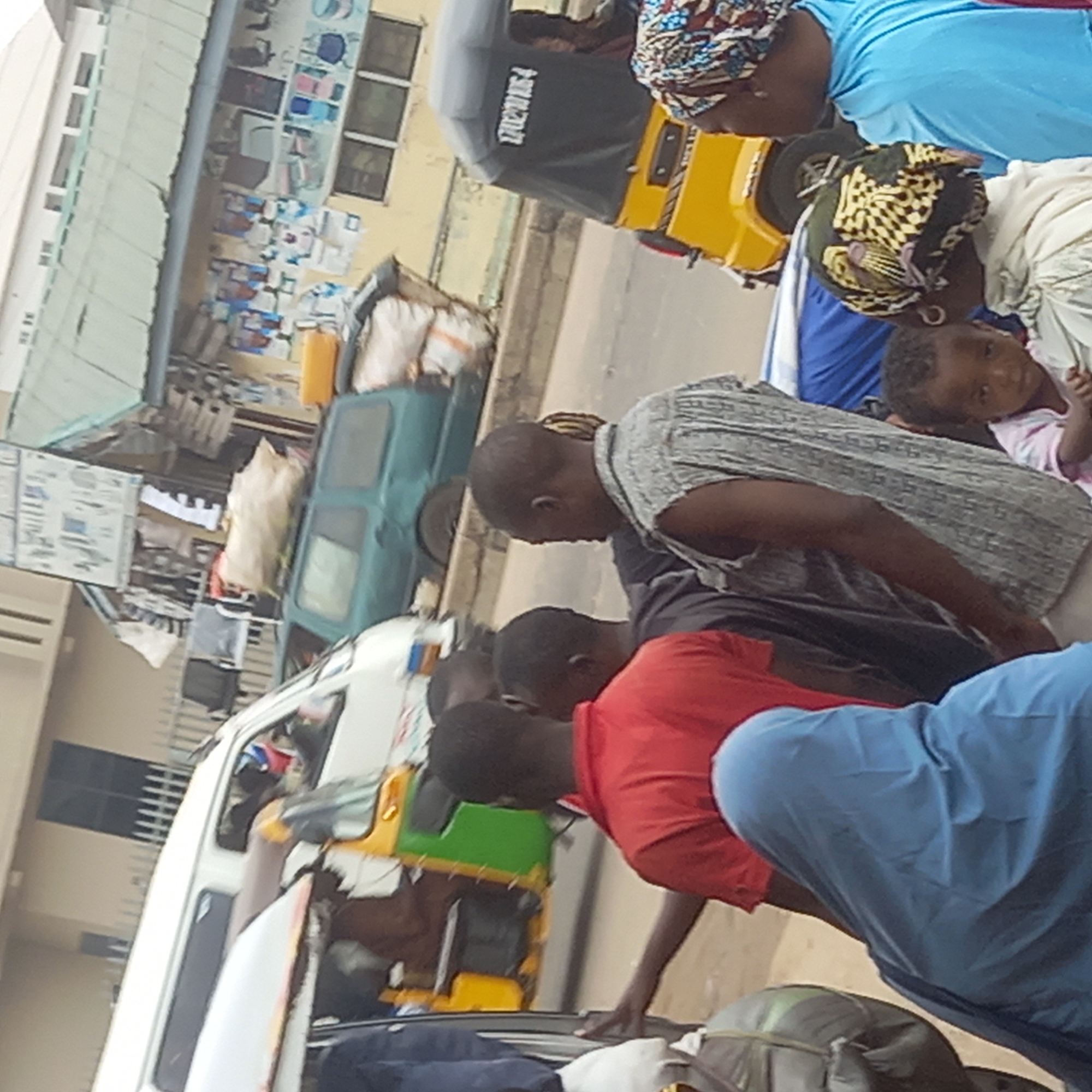 In Dadi Riba, Nigeria, the image shows individuals gathered near vehicles on a street. People are loading or unloading items. A yellow tricycle is visible. Buildings are present along the street. Luggage is on the ground.