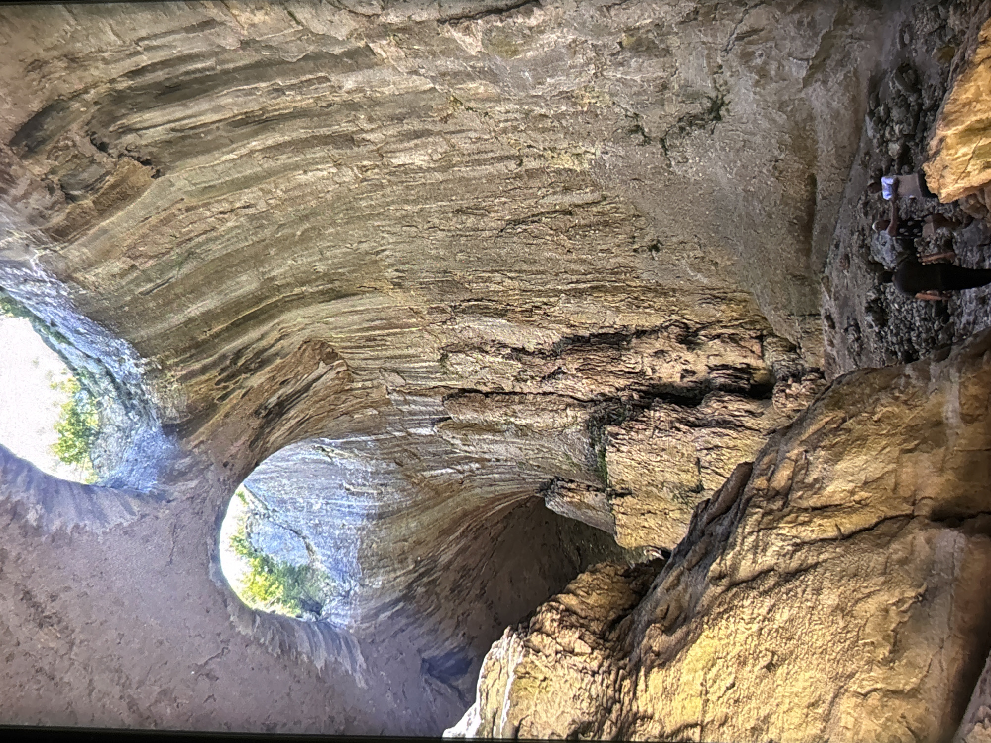 This image captures the impressive interior of a vast natural cave or rock formation, characterized by towering, layered rock walls and ceiling. The perspective looks upwards and across the cavern, revealing the intricate geological patterns formed by erosion and sedimentation, with textures ranging from smooth striations to rugged, cracked surfaces in shades of grey, brown, and ochre. Patches of green moss or algae cling to some areas of the rock.
On the left side of the frame, two large, circular openings allow bright daylight to penetrate the cave, offering glimpses of lush green foliage and trees outside, indicating a natural, open environment beyond. These openings suggest it is daytime. On the right, two people are visible, providing a sense of scale to the immense rock structure. One individual, wearing a light-colored shirt and dark pants, stands on a rocky ledge, while another person in dark clothing is positioned lower down, appearing to explore or observe their surroundings. The activity taking place is the exploration of this natural wonder.
The cavern's interior is illuminated by the natural light streaming through the openings, creating areas of bright light and deep shadow that highlight the contours and depth of the rock. There is no visible text in the image. While the provided location context is Wolverhampton, United Kingdom, the geological features depicted suggest a grand natural cave system.