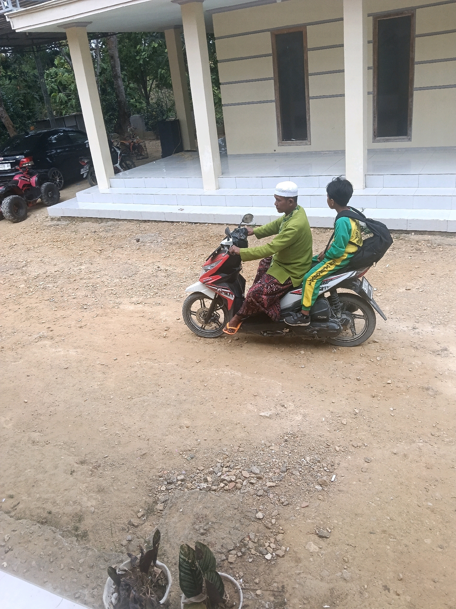 The image depicts a sunny outdoor scene on a dirt road in Bulangan Barat, Indonesia. In the mid-ground, a man wearing a light green long-sleeved shirt, a white cap, and a patterned sarong is riding a red and white scooter. A boy, dressed in a yellow and green long-sleeved shirt and matching pants, is seated behind the man, wearing a black backpack. They are both facing right, suggesting movement in that direction.
In the background, a building with a light-colored facade and dark windows stands. To the left of the building, a black car and a red all-terrain vehicle (ATV) are visible. The ground is covered in dirt and small stones, with a few potted plants in white containers in the foreground. The overall impression is a casual, everyday scene in a rural or semi-rural Indonesian setting.