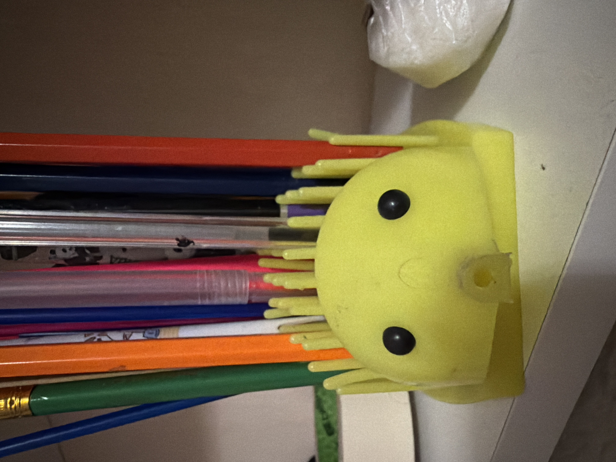 A bright yellow toy, resembling a creature with two black eyes and a gaping mouth, sits on a shelf. The toy has several yellow protrusions sticking out from its top, resembling spikes or hair. Behind the toy, a collection of colorful pens and pencils stand upright in a container, some with gold ferrules. The pens and pencils are of various colors, including blue, green, orange, white, purple, pink, and red. The shelf appears to be made of a light-colored material, possibly wood or laminate. To the right of the toy, a white object wrapped in plastic is visible. The background is a plain, textured wall of a light brown color. There are no people visible in the image. The scene appears to be indoors, likely in a home or office setting. There is no information in the image that would allow for determination of the location in the city of Ulan-Ude, Russia.