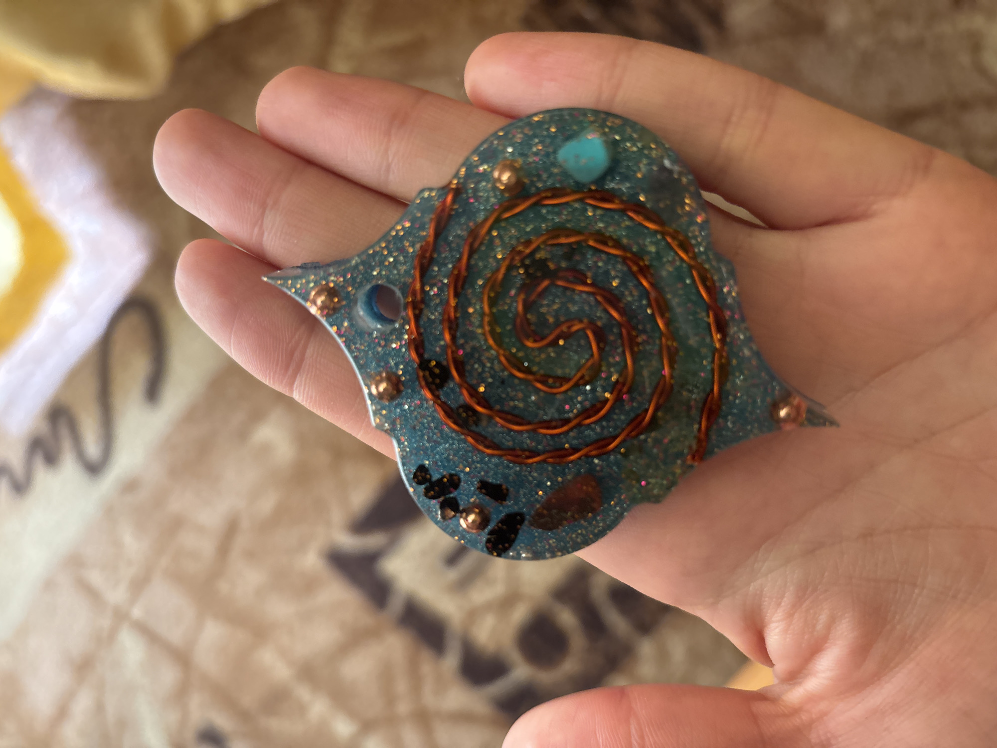 A person's hand holds a blue, glittery resin pendant shaped like an elongated hexagon with two points extending from one side. The pendant is embedded with various objects, including small dark stones, copper-colored beads, a spiral of copper wire, and a small turquoise stone. The background is a blurred, patterned fabric in shades of brown and yellow.