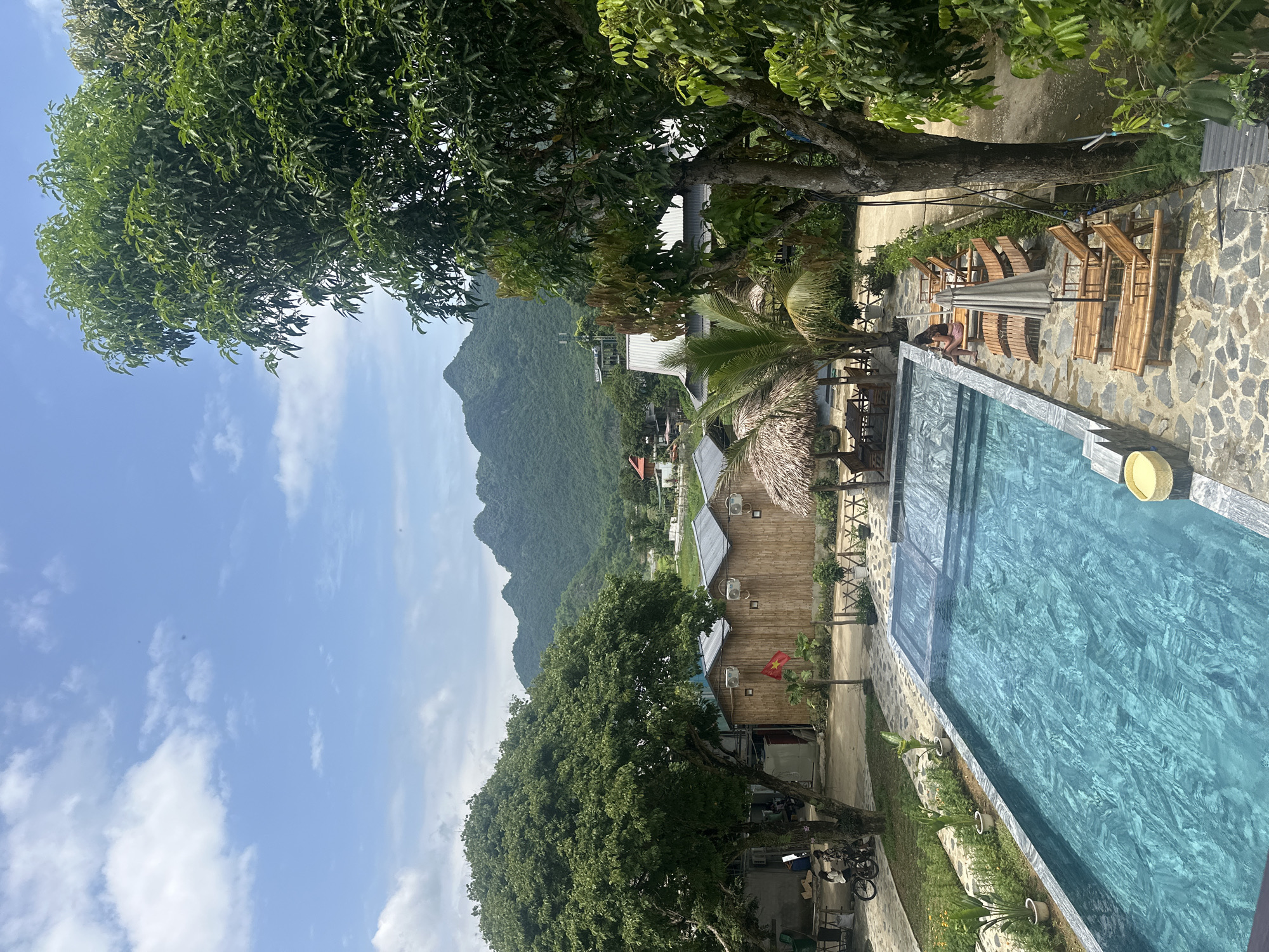 This is a medium-angle, eye-level shot of a resort area in Xóm Khán, Vietnam, on a partly cloudy day. In the foreground, a rectangular swimming pool with turquoise water is set against a stone patio. Several wooden lounge chairs are arranged around the pool, with one person seated in a chair on the far side.
To the left of the pool, a grassy area is bordered by plants and a paved walkway. A tree stands prominently on the left, with a parked motorcycle and two people standing beside it.
In the middle ground, several small, bamboo-constructed cabins with thatched roofs are visible, along with a palm tree and a covered seating area with tables. A Vietnamese flag is displayed on a pole in front of one of the cabins.
The background features rolling, lush green mountains under a blue sky with scattered white clouds. The overall scene depicts a tranquil resort setting amidst a natural landscape.