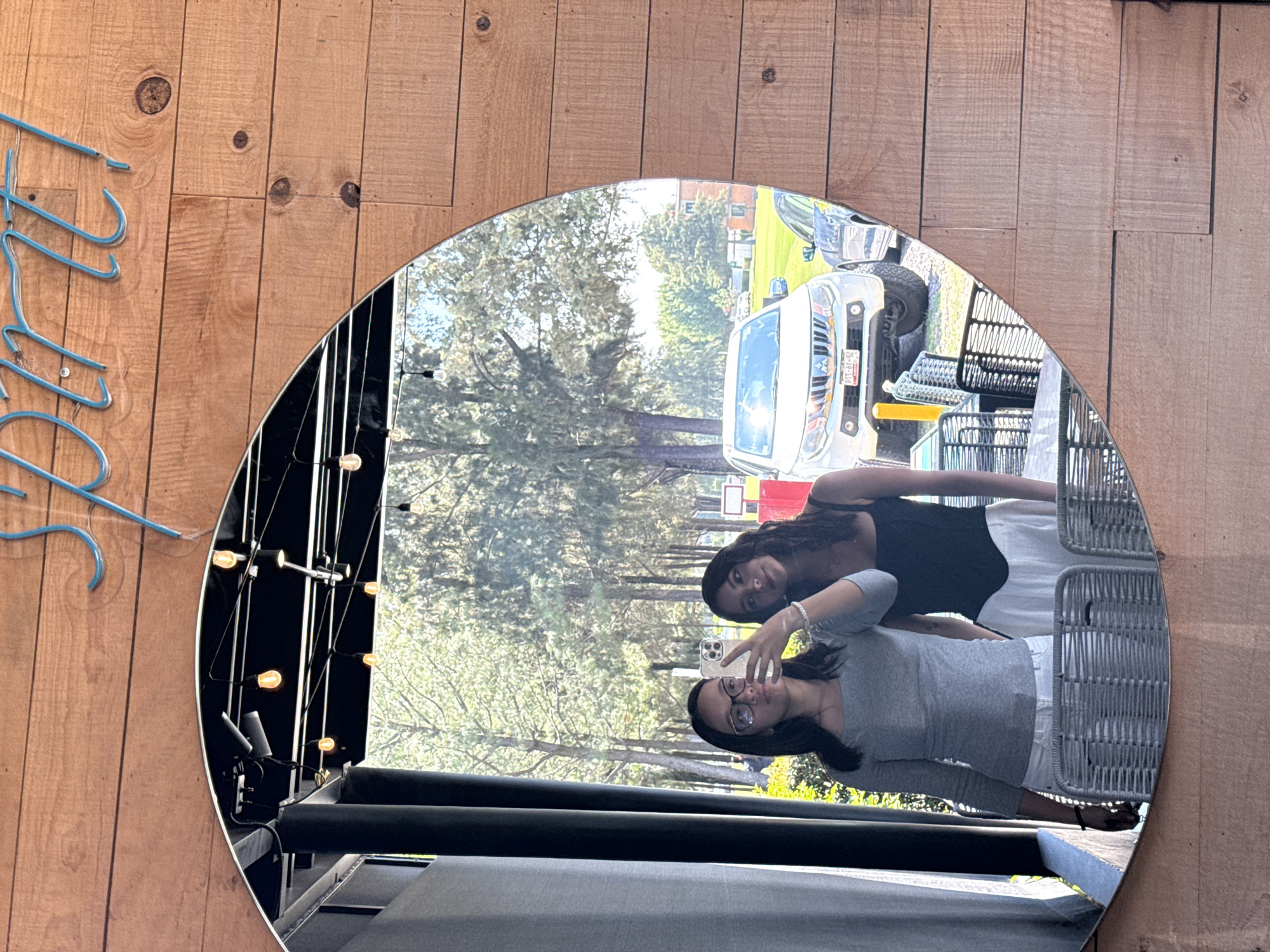 The image shows a selfie of two women reflected in a circular mirror, located in San Andrés Cholula, Mexico. The reflection captures the women, a light-colored truck with license plate "ATC-07-92" and the exterior of a building. The woman in the foreground is wearing a gray shirt, glasses, and pointing at the camera. The second woman, positioned behind her, is wearing a black top. The mirror is mounted on a wooden wall next to a light blue neon sign. The reflection also reveals outdoor furniture and greenery. Inside the space behind the mirror, there is a series of lights hanging from black fixtures.